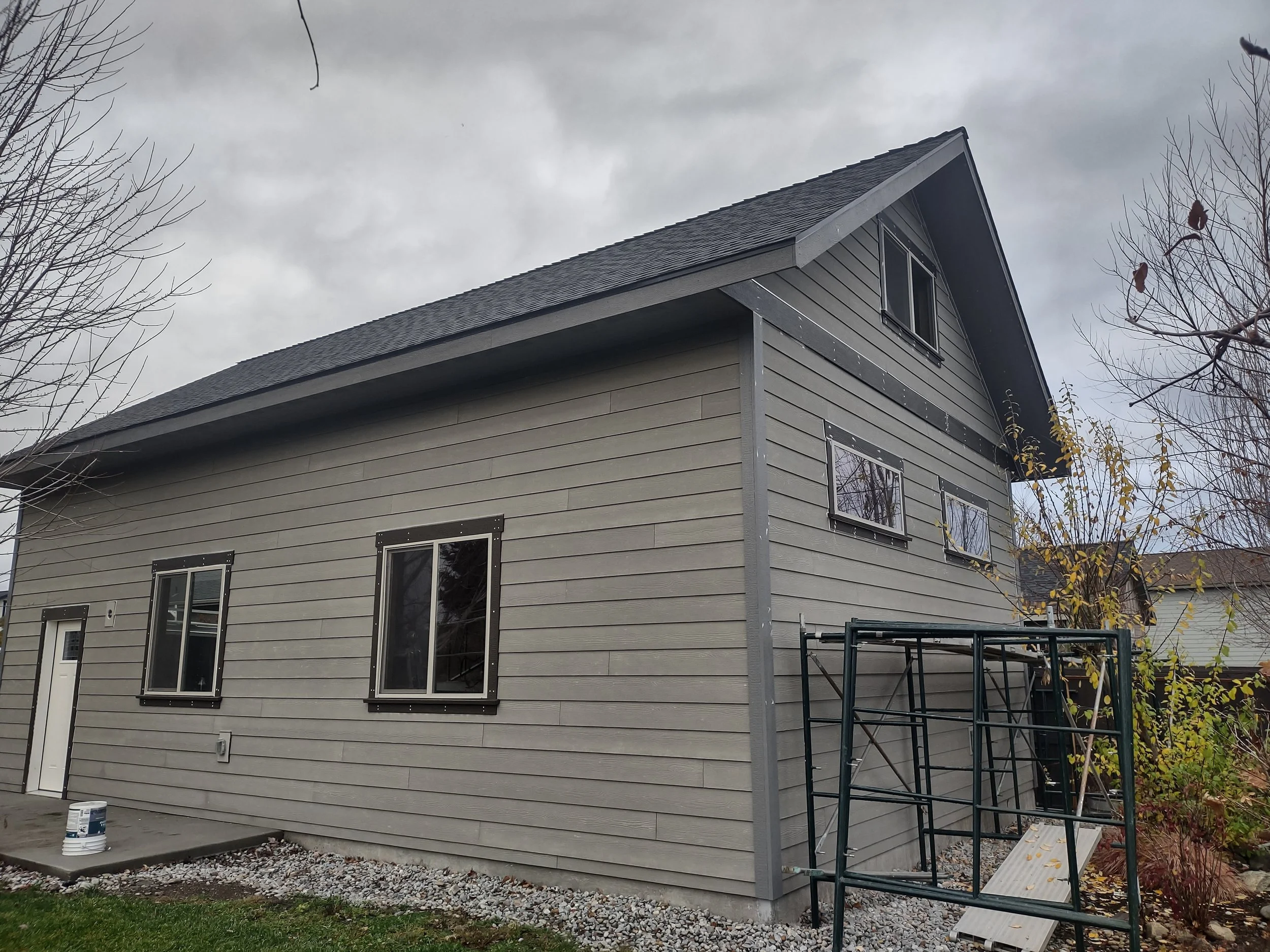 A two-story house with gray horizontal siding, several windows with black framing, and a steep gabled roof. There are leafless trees and a scaffold outside, with a cloudy sky overhead.