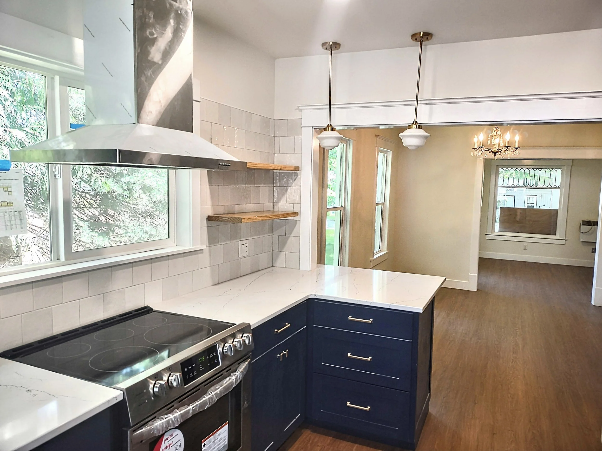 Modern kitchen with white marble countertops, blue cabinets, stainless steel oven, white tiled backsplash, wooden open shelves, and a range hood. Adjacent dining area features wood flooring, windows, and a chandelier.