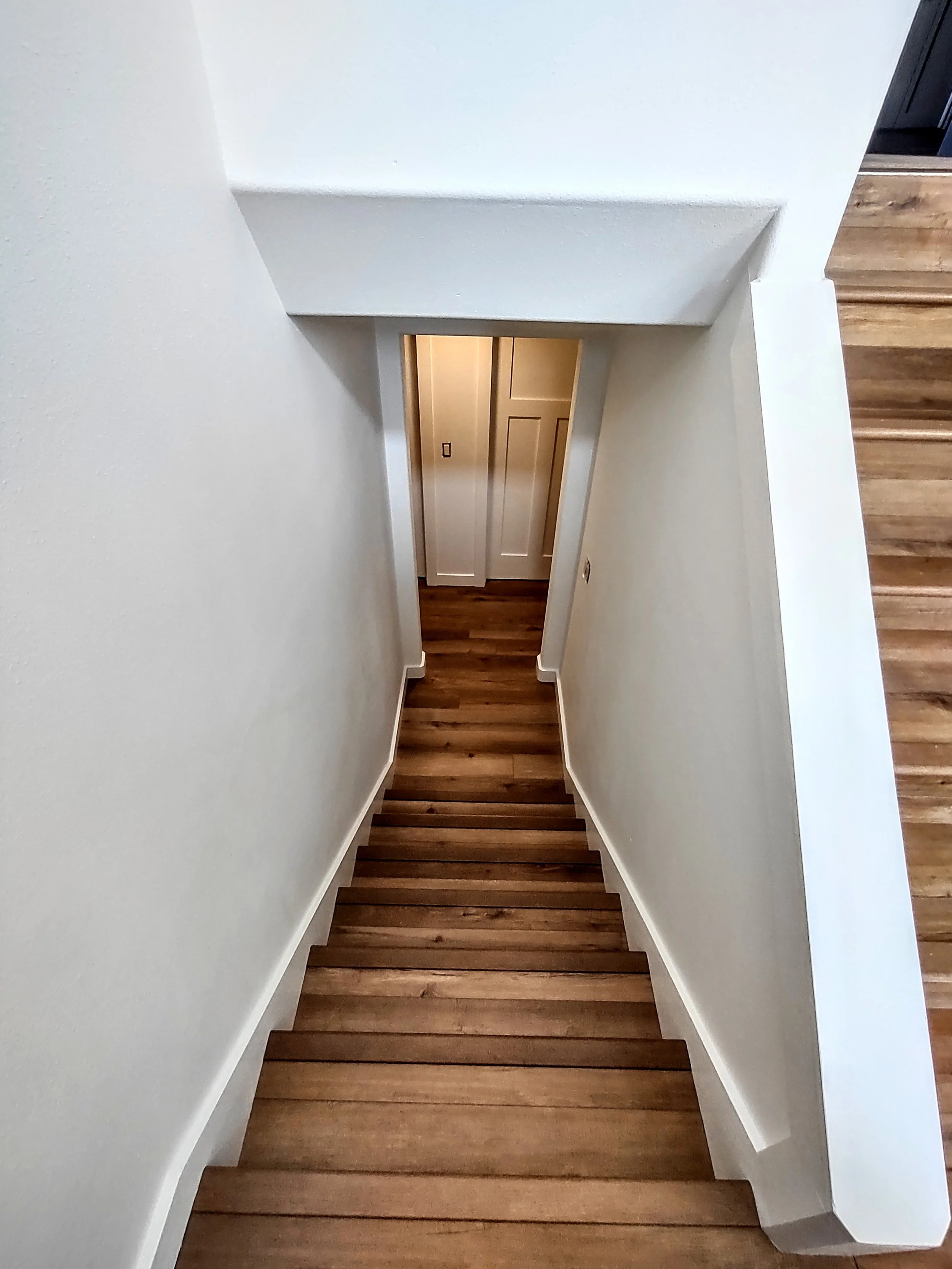 View looking down a staircase with wooden steps, white walls, and a door at the bottom.