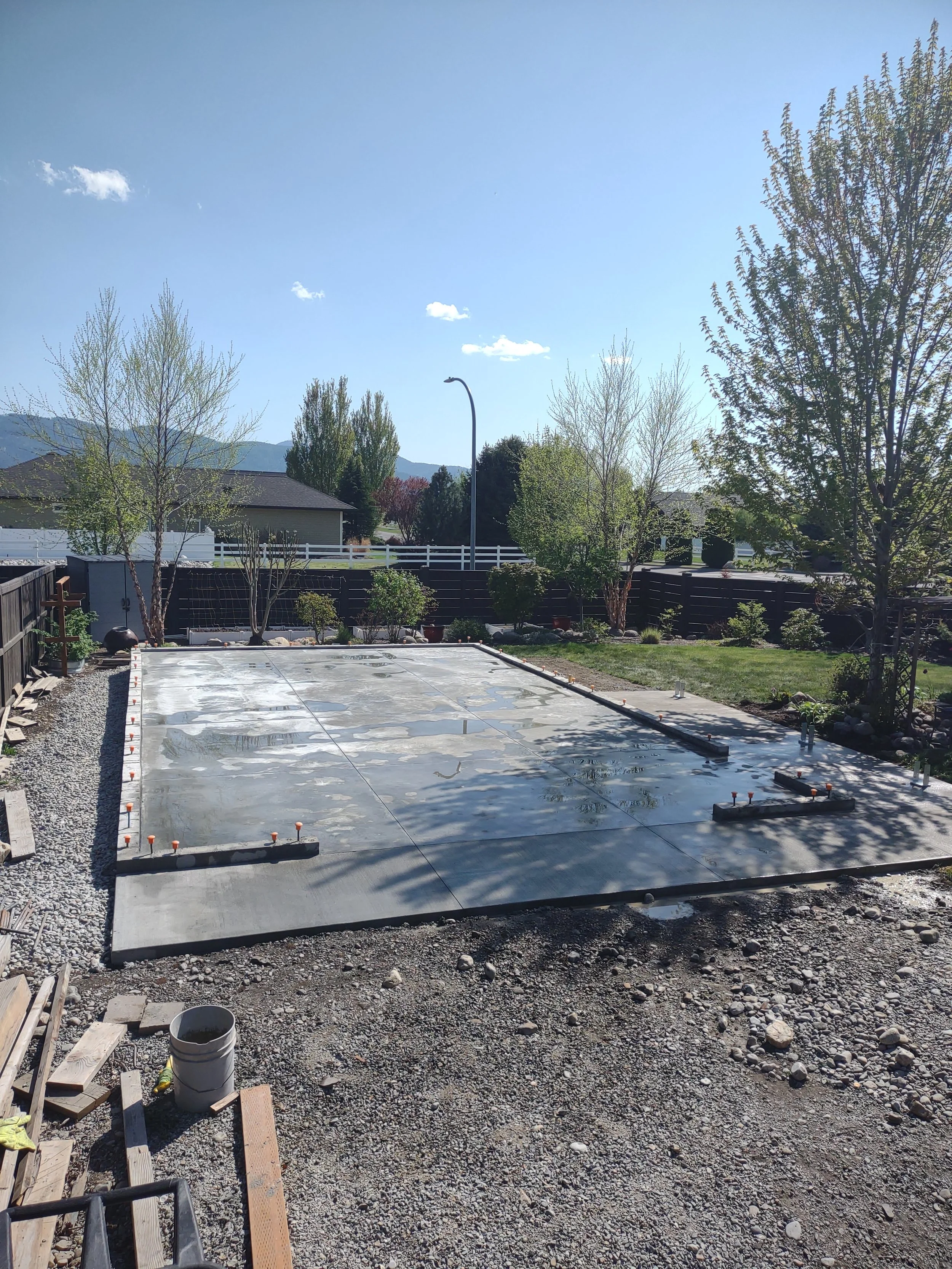 A backyard with a concrete foundation, surrounded by gravel, wood, and plants, with some trees in the background and a clear blue sky.