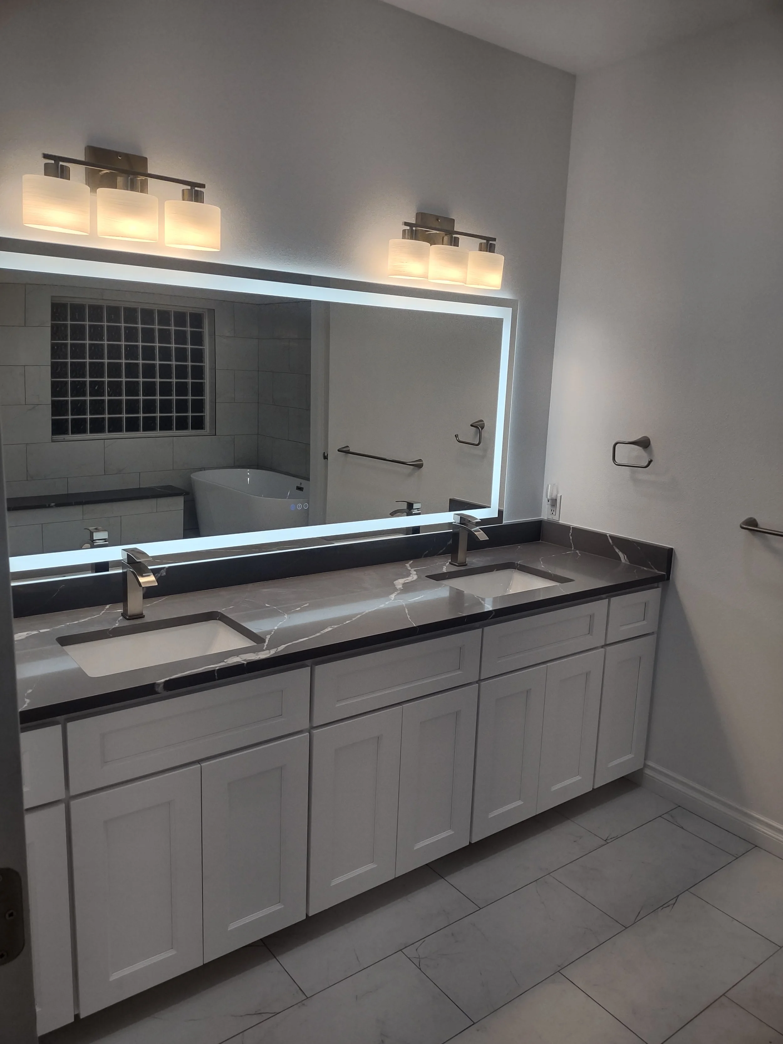 Bathroom with double vanity, marble countertop, and illuminated mirror. Wall-mounted light fixtures and chrome faucets. Reflected in mirror is bathtub, window, towel bars, and tiled wall.
