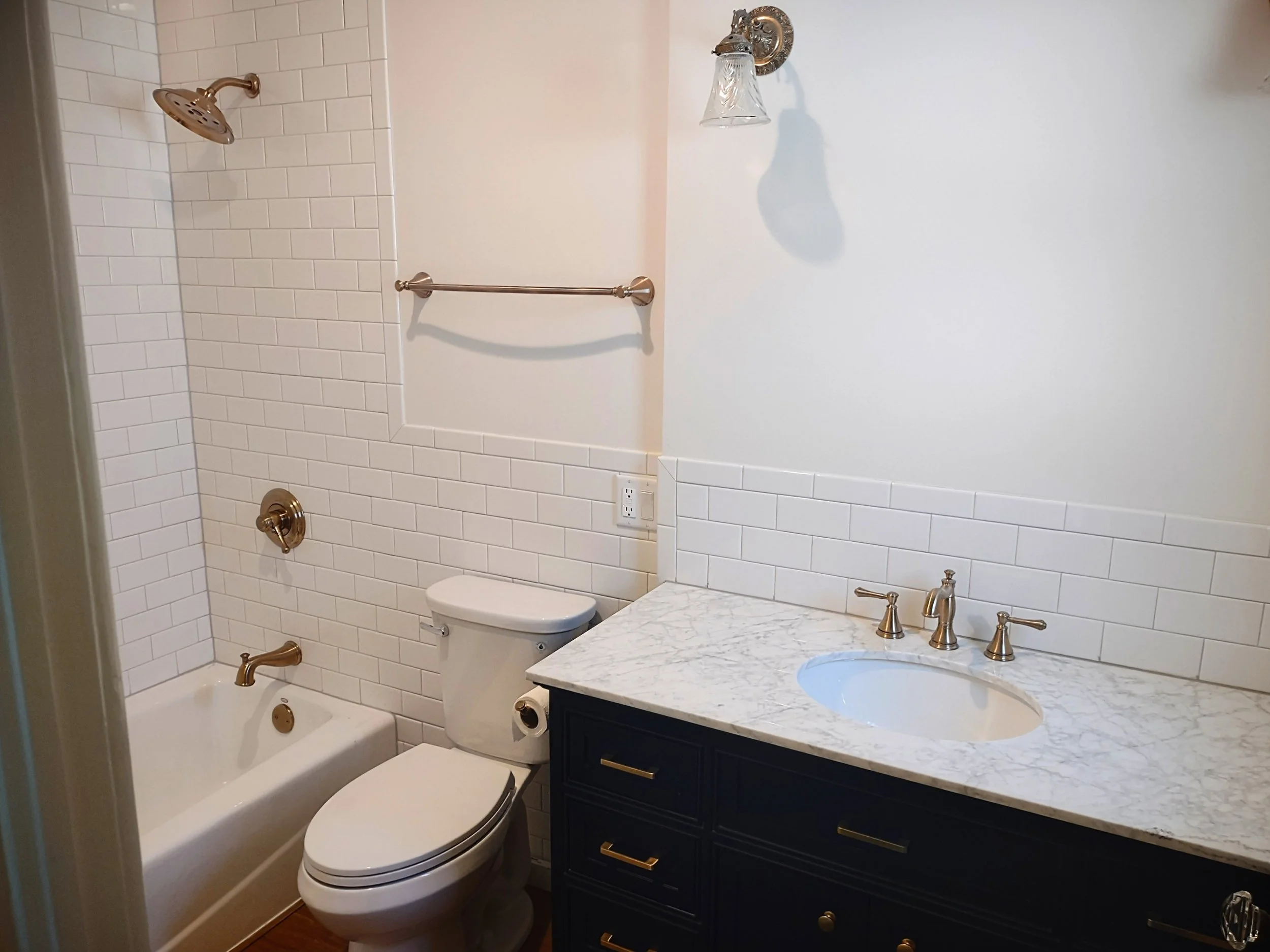 Bathroom with white subway tile walls, a bathtub with a gold faucet, a toilet, a dark blue vanity with a white marble countertop, a sink with gold fixtures, a towel bar, and a wall-mounted light fixture.