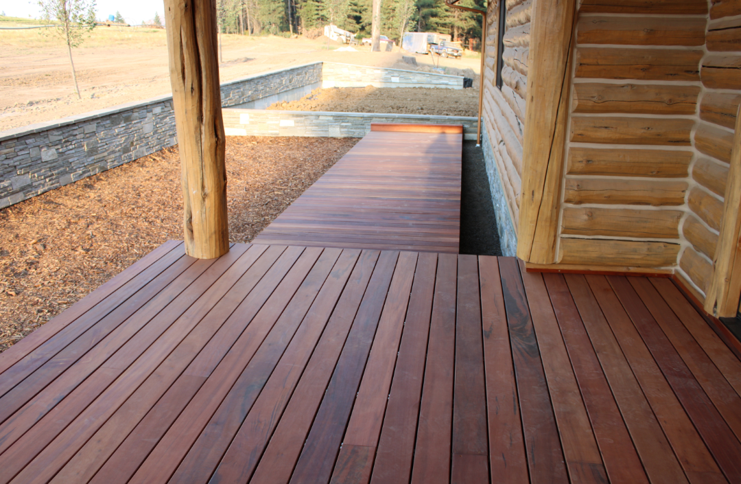 View of a wooden porch with a new deck made of stained wood, attached to a log cabin, with dirt yard and trees in the background.