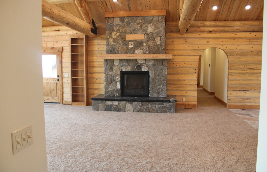 Living room with a stone fireplace, wooden walls, ceiling beams, and carpeting.