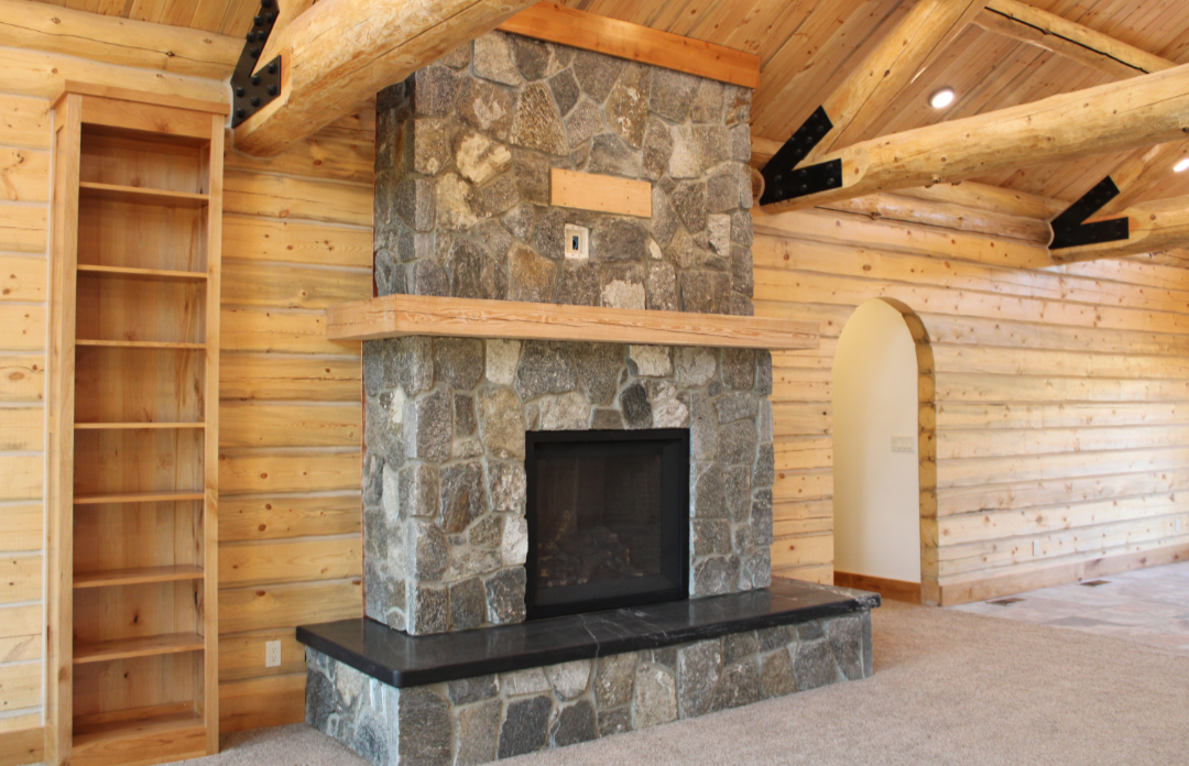 Interior of a wood cabin living room with a stone fireplace, wooden shelves, wood-paneled walls, and a wooden beam ceiling.