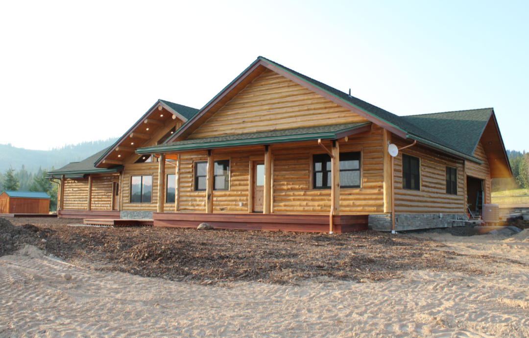 A large wooden house under construction with a green roof, set against a mountainous background and a clear sky.