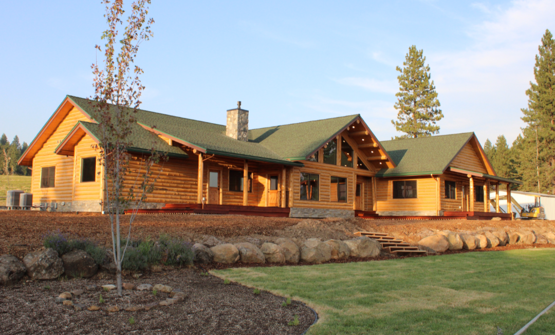 A large wooden house with a green roof, set in a landscaped yard with grass, rocks, and trees.