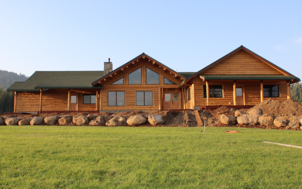 Wooden house under construction in a grassy yard with rocks along its foundation and a mountain in the background.