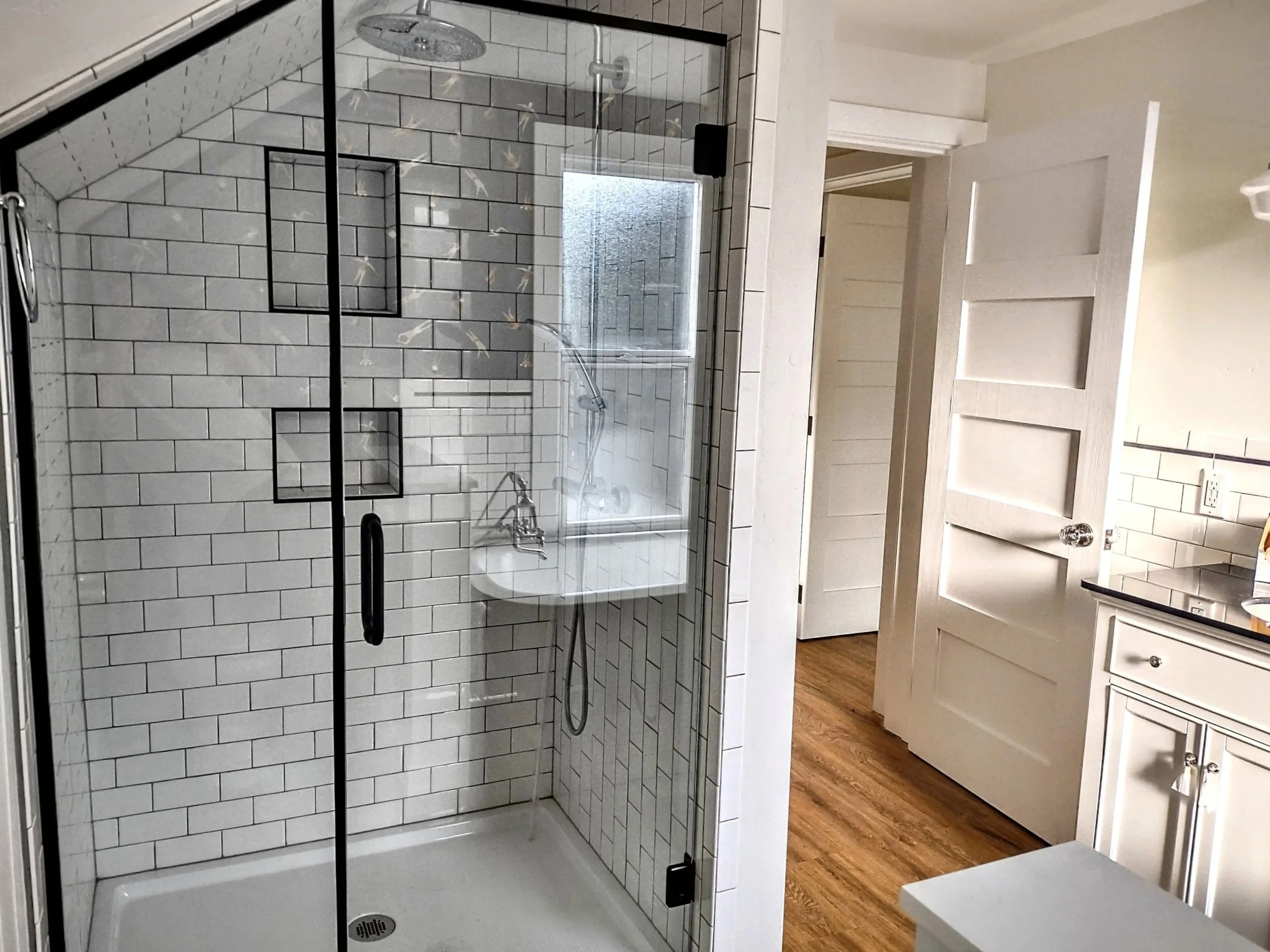 Bathroom with glass-enclosed shower featuring white subway tiles, black accent tiles, a rain shower head, and a handheld shower. To the right, there is a white wooden door, a cabinet with black countertop, and a wood floor.