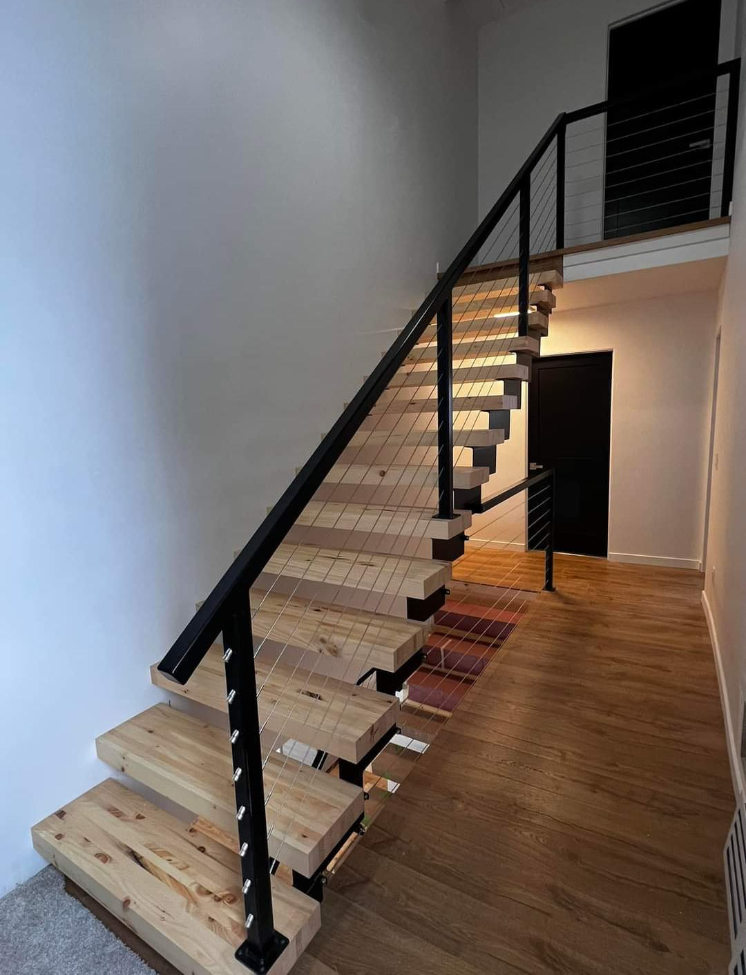 Interior view of a modern staircase with wooden steps and black metal railing in a well-lit room with wooden flooring.