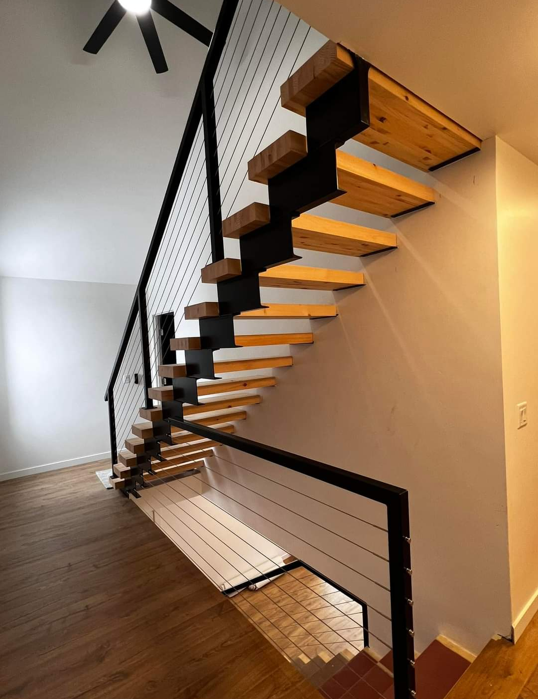 Interior view of a modern open-riser staircase with wooden treads and black metal supports, featuring cable railings, in a room with wood flooring and white walls.