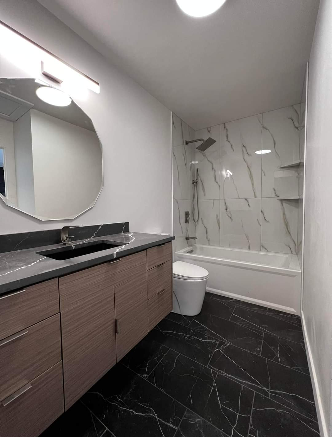 Modern bathroom with black marble floor, brown vanity with black marble countertop, white sink, large mirror, and a bathtub with marble wall tiles and a showerhead.