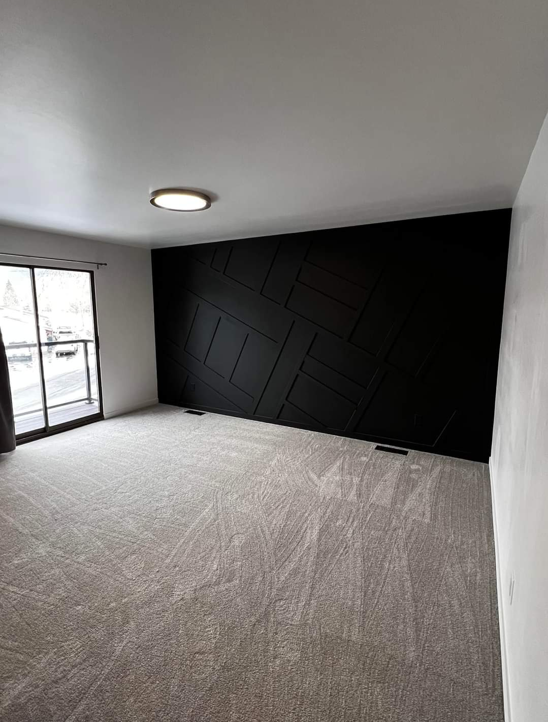 Empty room with beige carpet, white walls, and a black accent wall with geometric panel design. Large glass sliding door leads to a balcony, with natural light coming in and a ceiling light fixture installed.