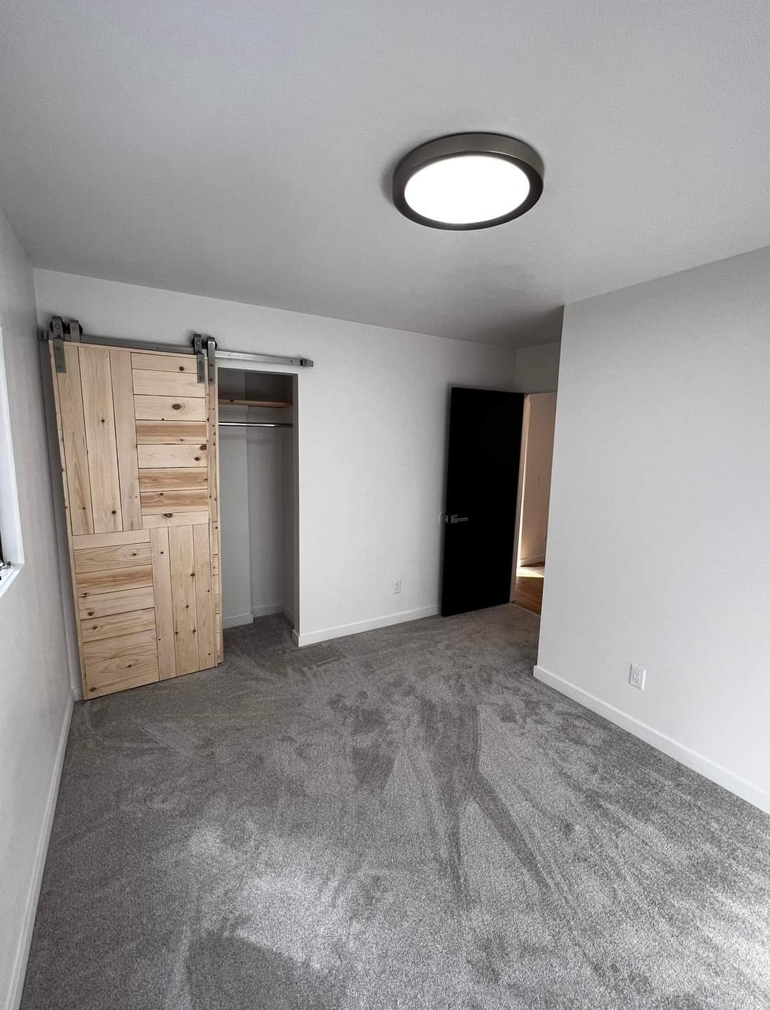 Empty bedroom with grey carpet, white walls, a black door, an open closet with sliding wooden doors, and a ceiling light.