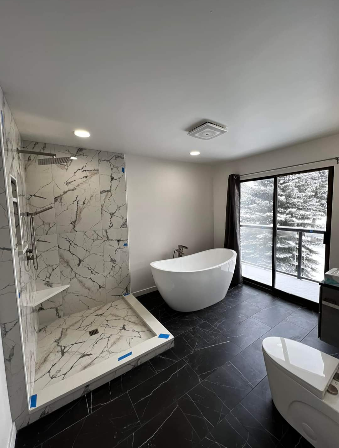 Modern bathroom with a white bathtub, a walk-in shower with marble tiles, a toilet, and a large window showing trees outside.