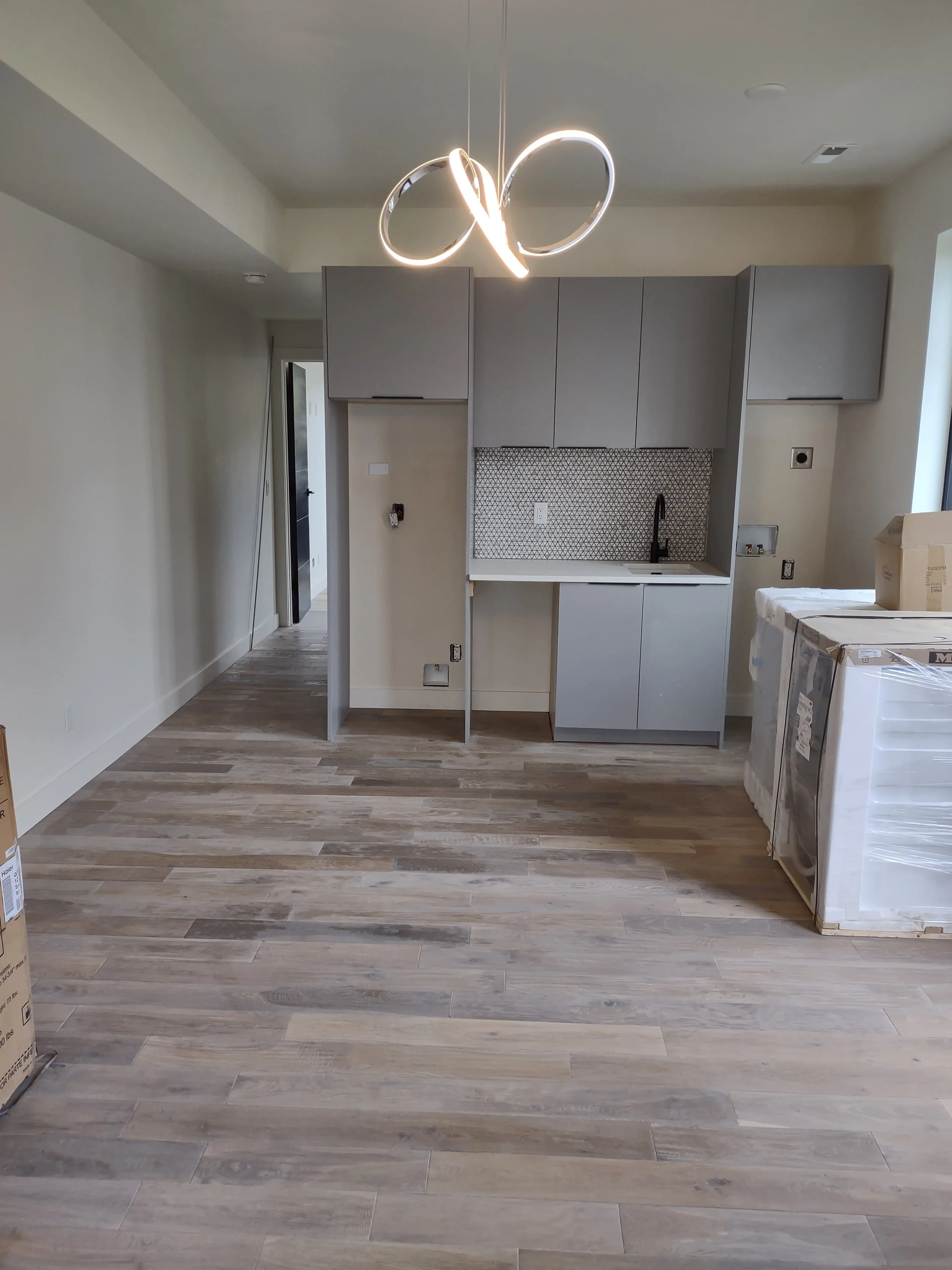 Empty kitchen with light wood flooring, gray cabinets, and a geometric backsplash above the sink. Overhead modern light fixture with intertwined rings. Packaging on the right side, partially enclosed.