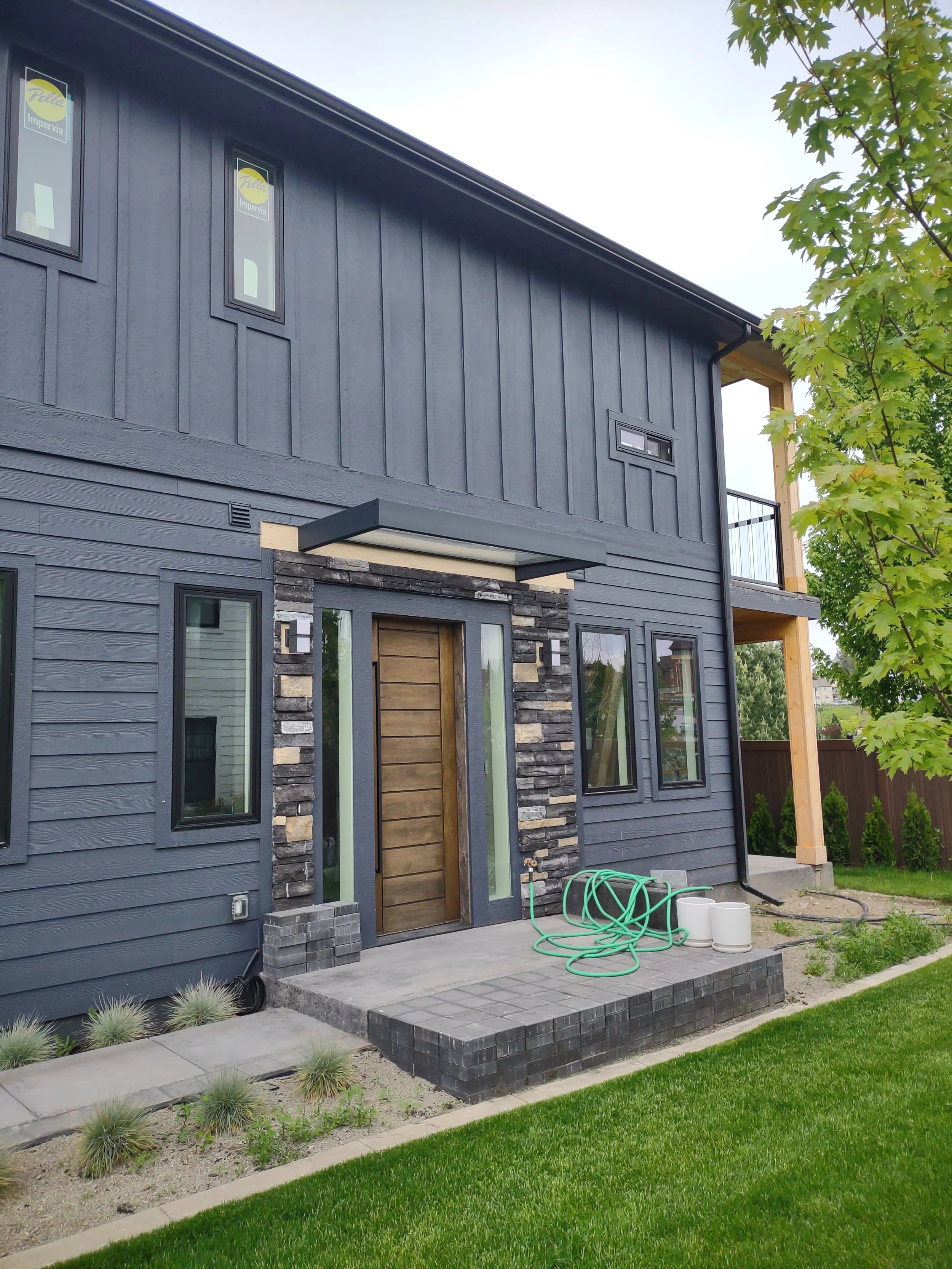 A modern two-story house with dark gray siding, a wooden front door, and a small front porch with landscaping and a garden hose coiled on the ground.