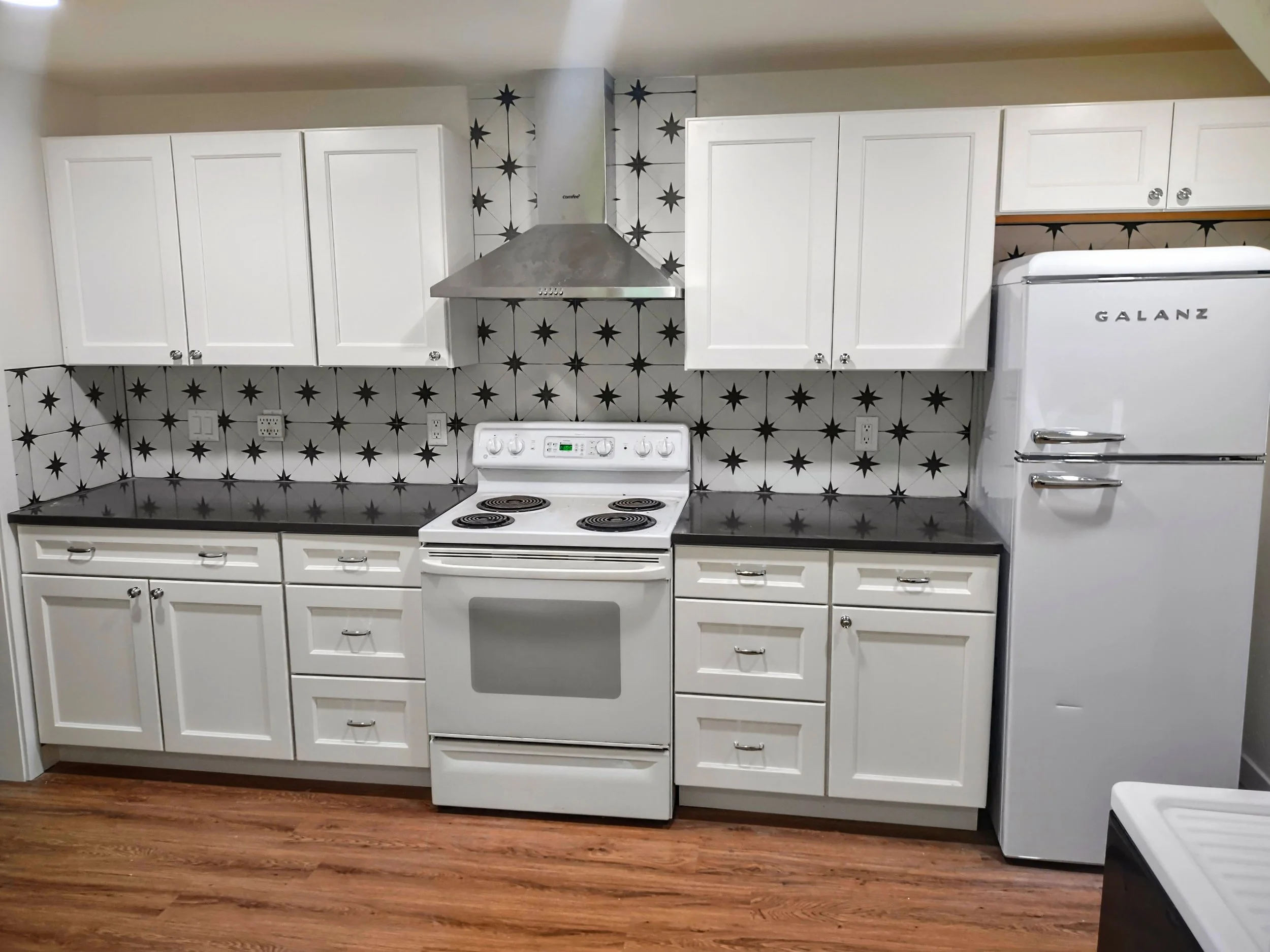 Kitchen with white cabinets, black countertop, white stove, silver range hood, Galanz refrigerator, and black-and-white star-patterned tile backsplash on wood floor.