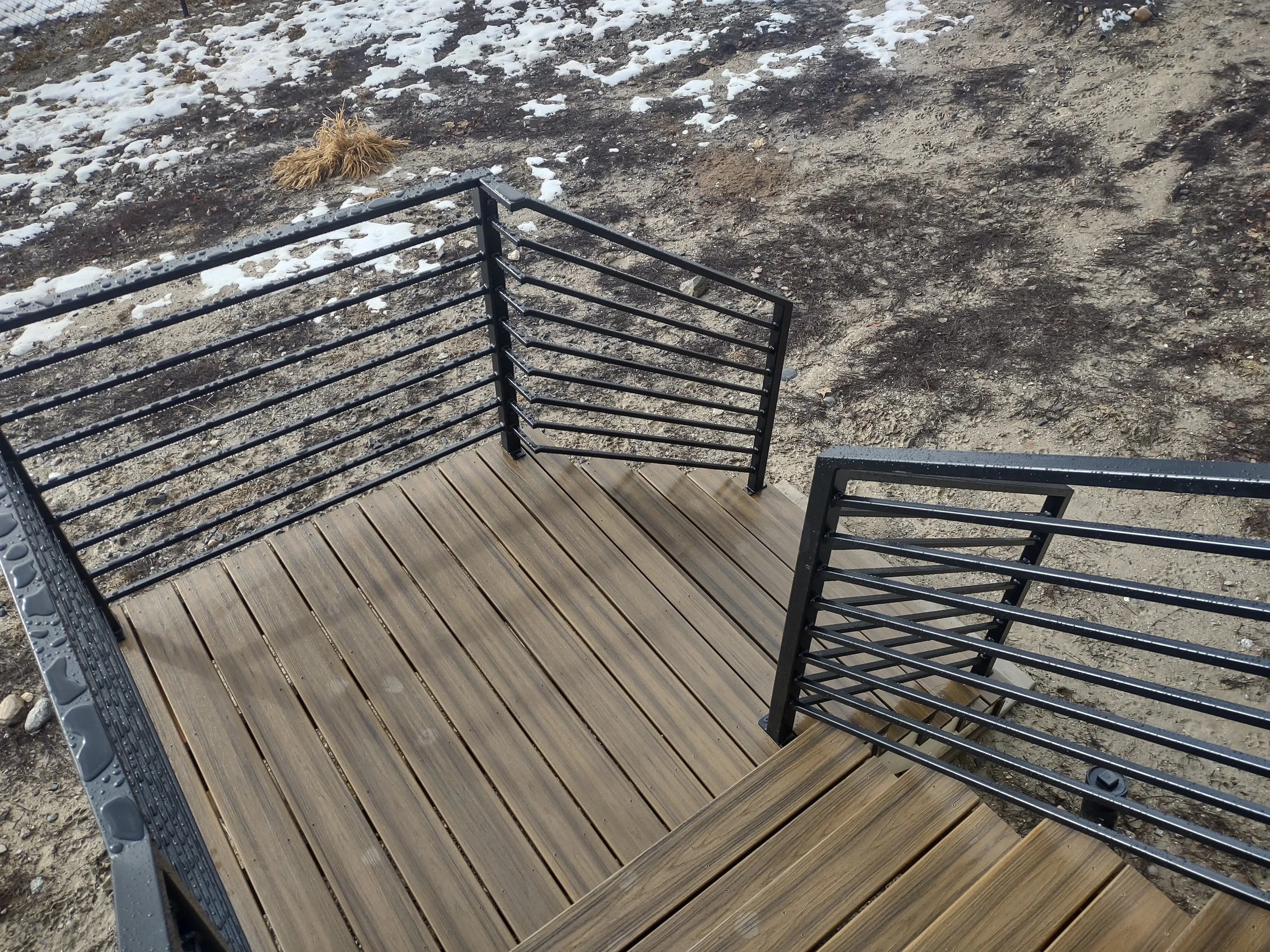 Bare wooden deck with black metal railing on dirt ground, patches of snow, and a small section of dried grass