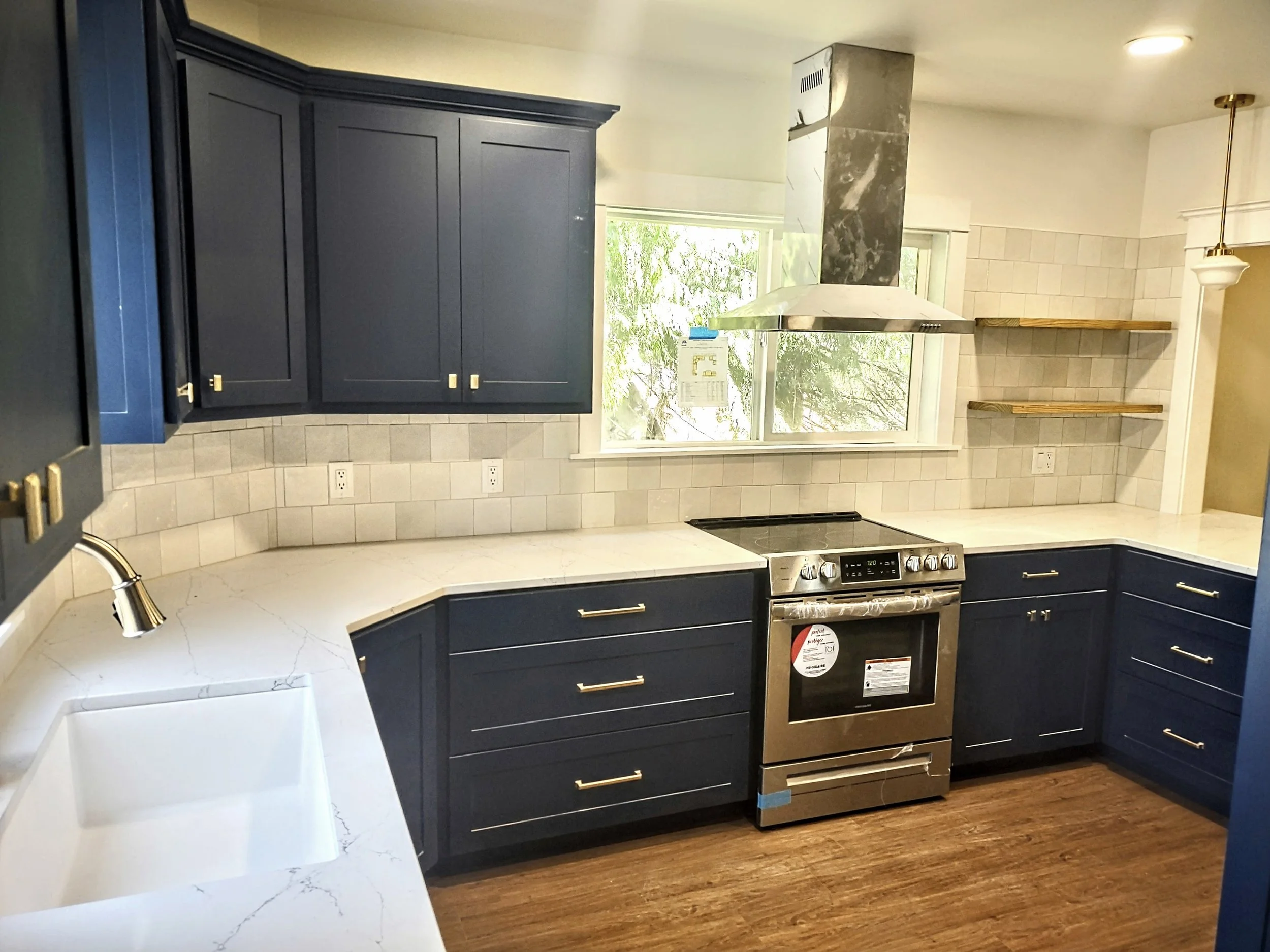 A modern kitchen with dark blue cabinets, a marble countertop, a stainless steel stove, a white farmhouse sink, a window above the stove, a range hood, and wooden open shelves on a tiled wall.