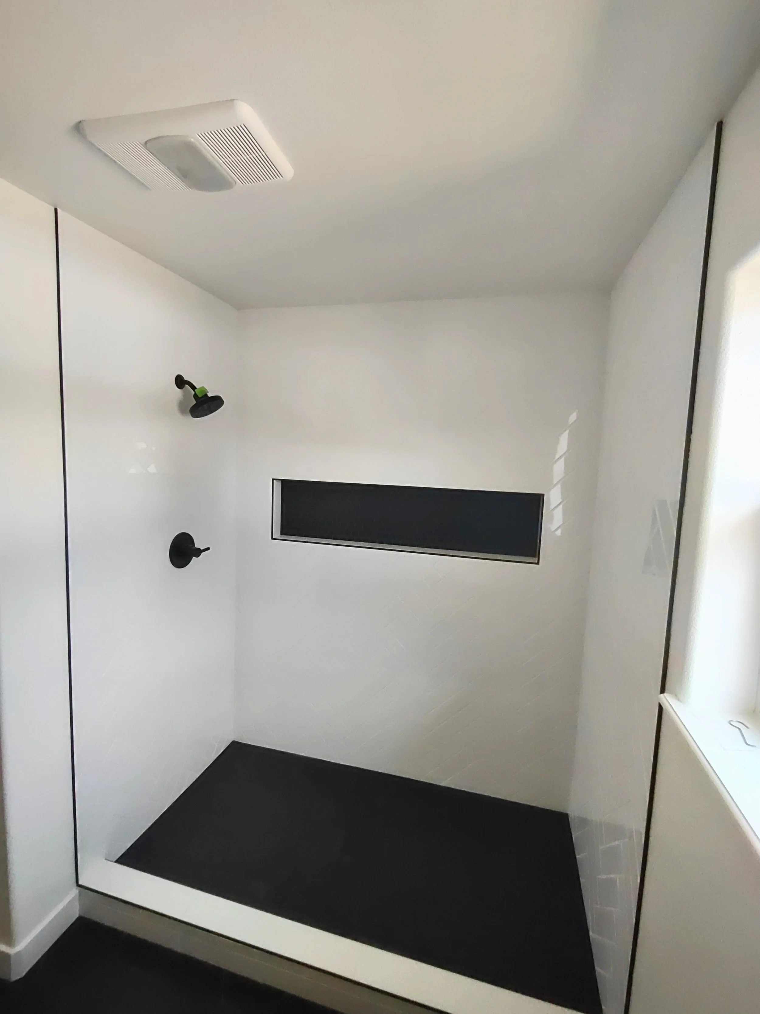 Empty walk-in shower with black fixtures, a small black horizontal window, and a white tiled wall in a modern bathroom.
