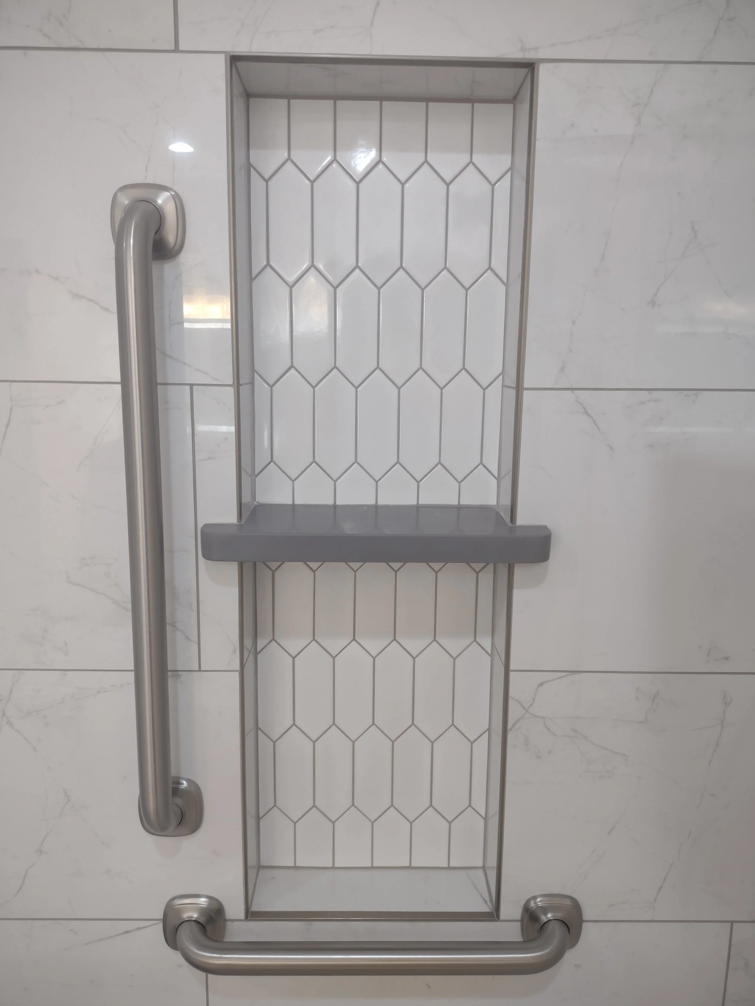 A shower niche with white hexagon tile, two gray shelves, and stainless steel grab bars on a white marble tiled wall.