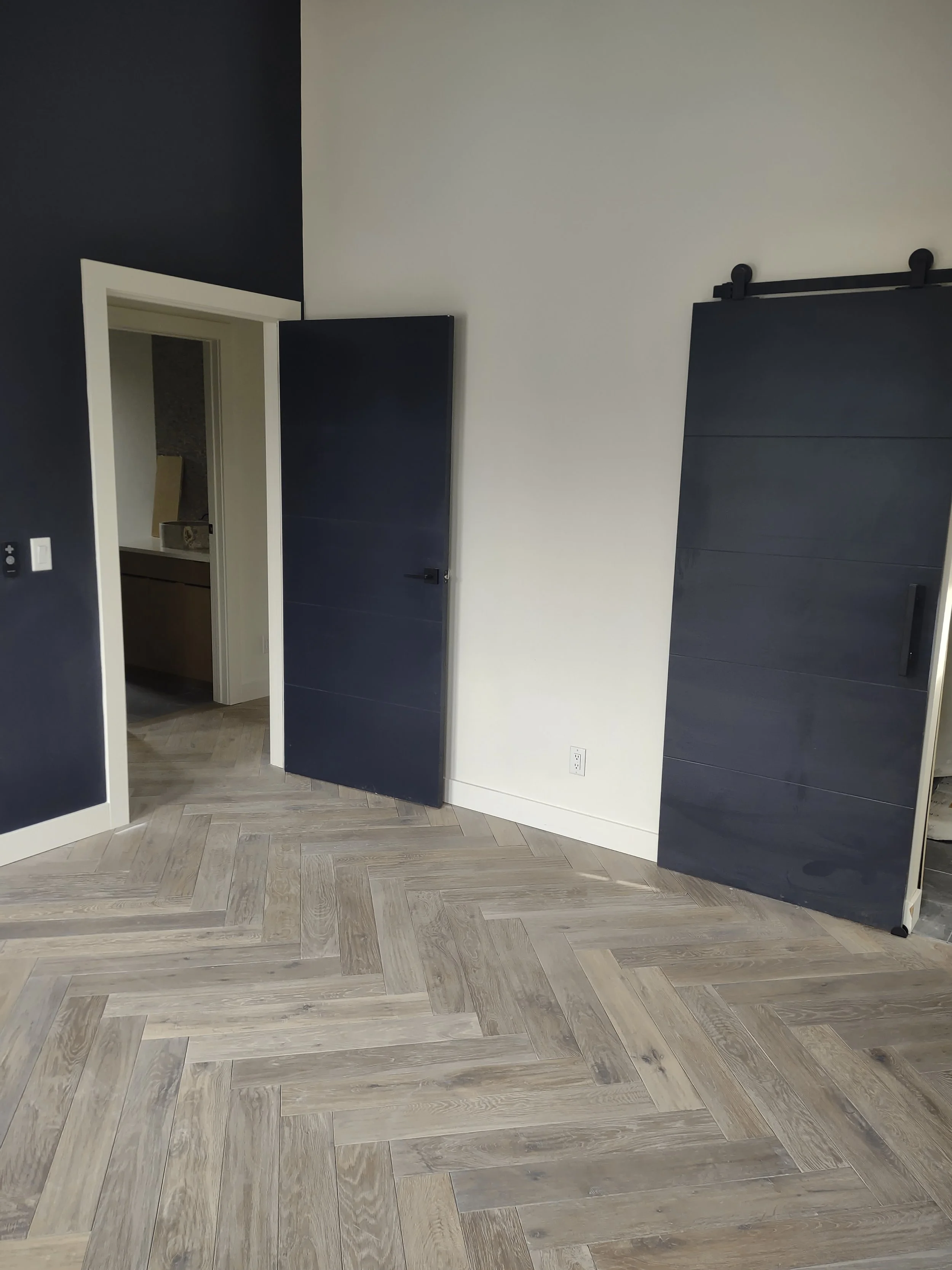 Empty room with wooden herringbone floor, dark and light walls, two dark sliding barn doors, one open, leading to another room.