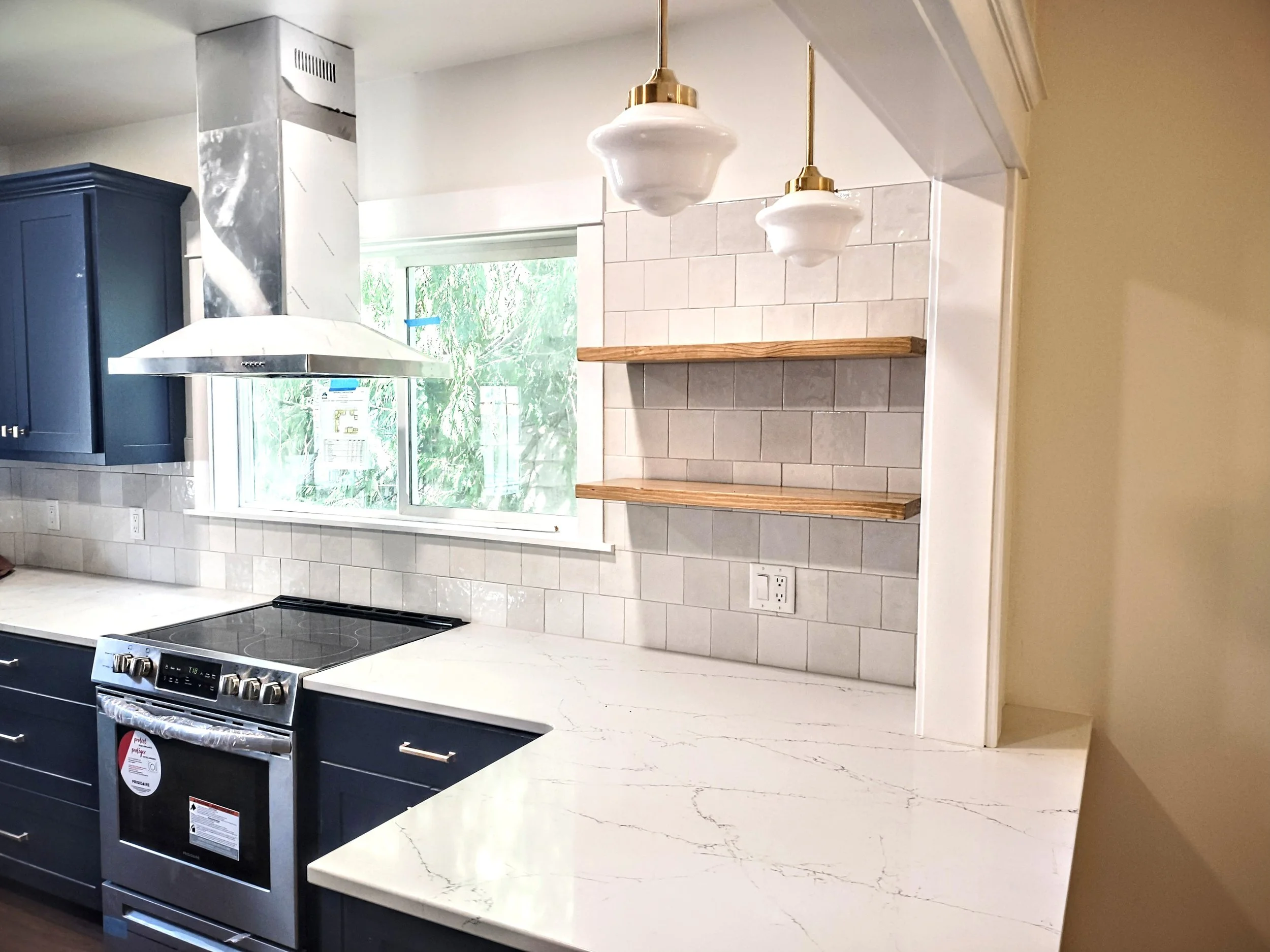 Modern kitchen with white marble countertop, blue cabinets, stainless steel stove, white tile backsplash, and a window showing greenery outside. Two white pendant lights hanging from the ceiling and open wooden shelves on the wall.