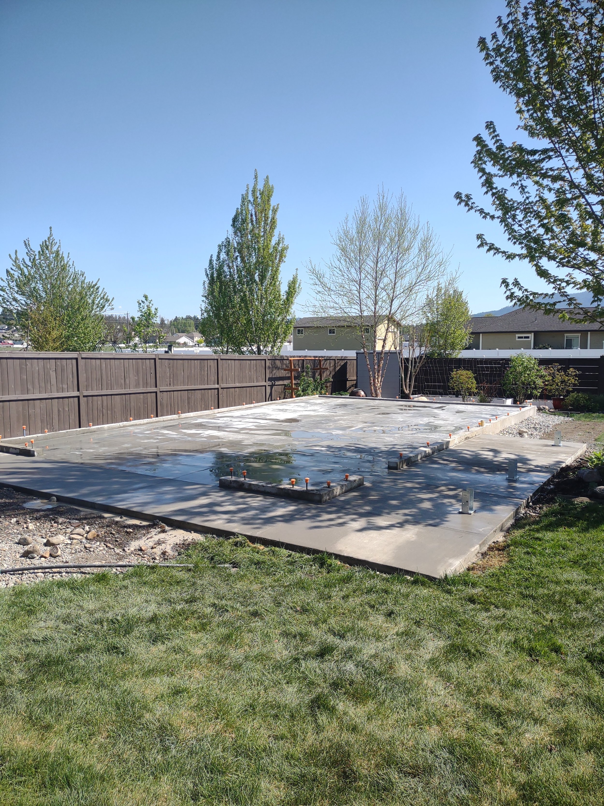 Constructed concrete foundation for a new building in a backyard, with a wooden fence and trees in the background, and a clear blue sky above.