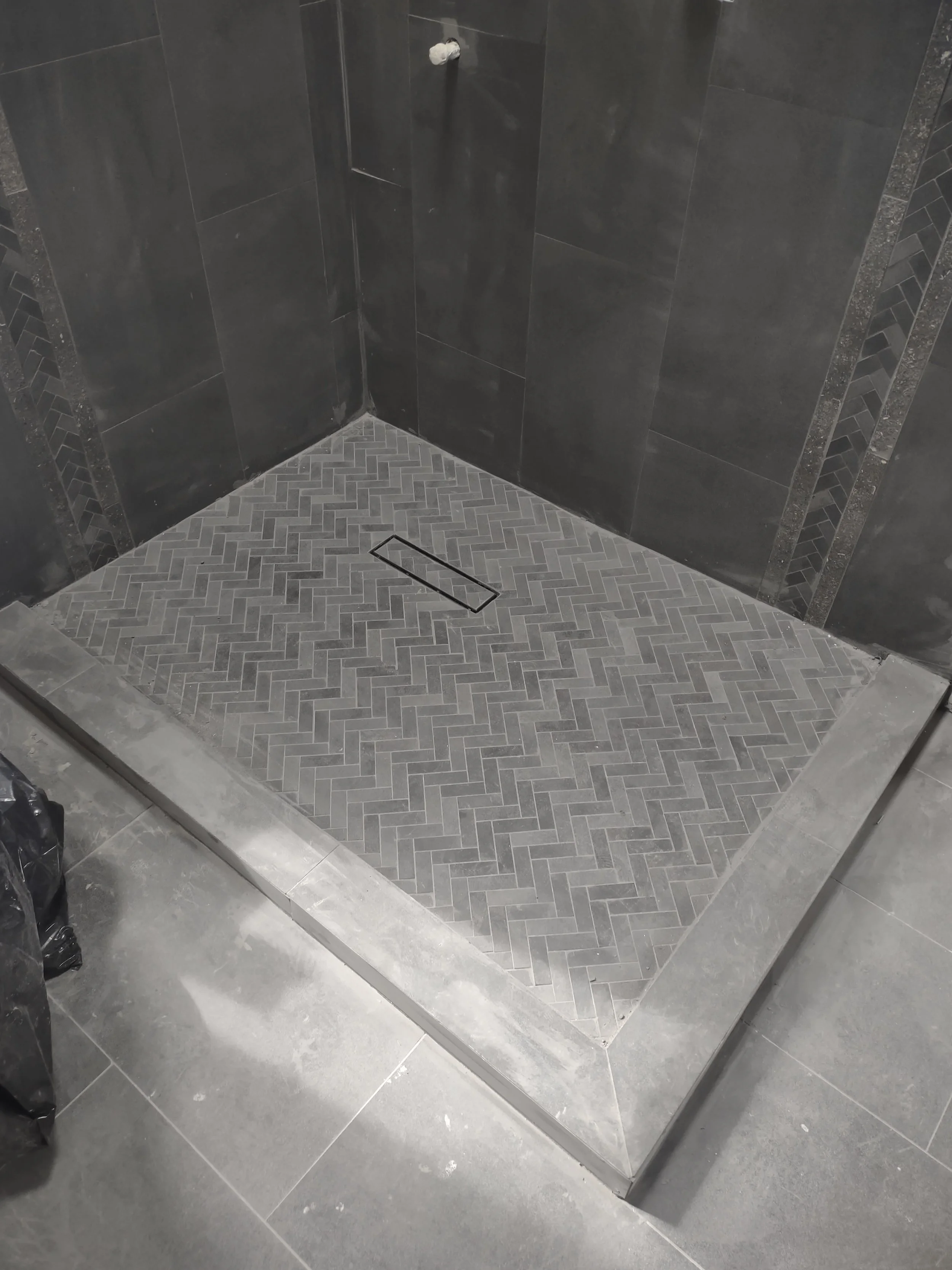 A shower stall with dark gray tiled walls, a herringbone patterned gray tiled floor, a linear drain, and a surrounding metal threshold.