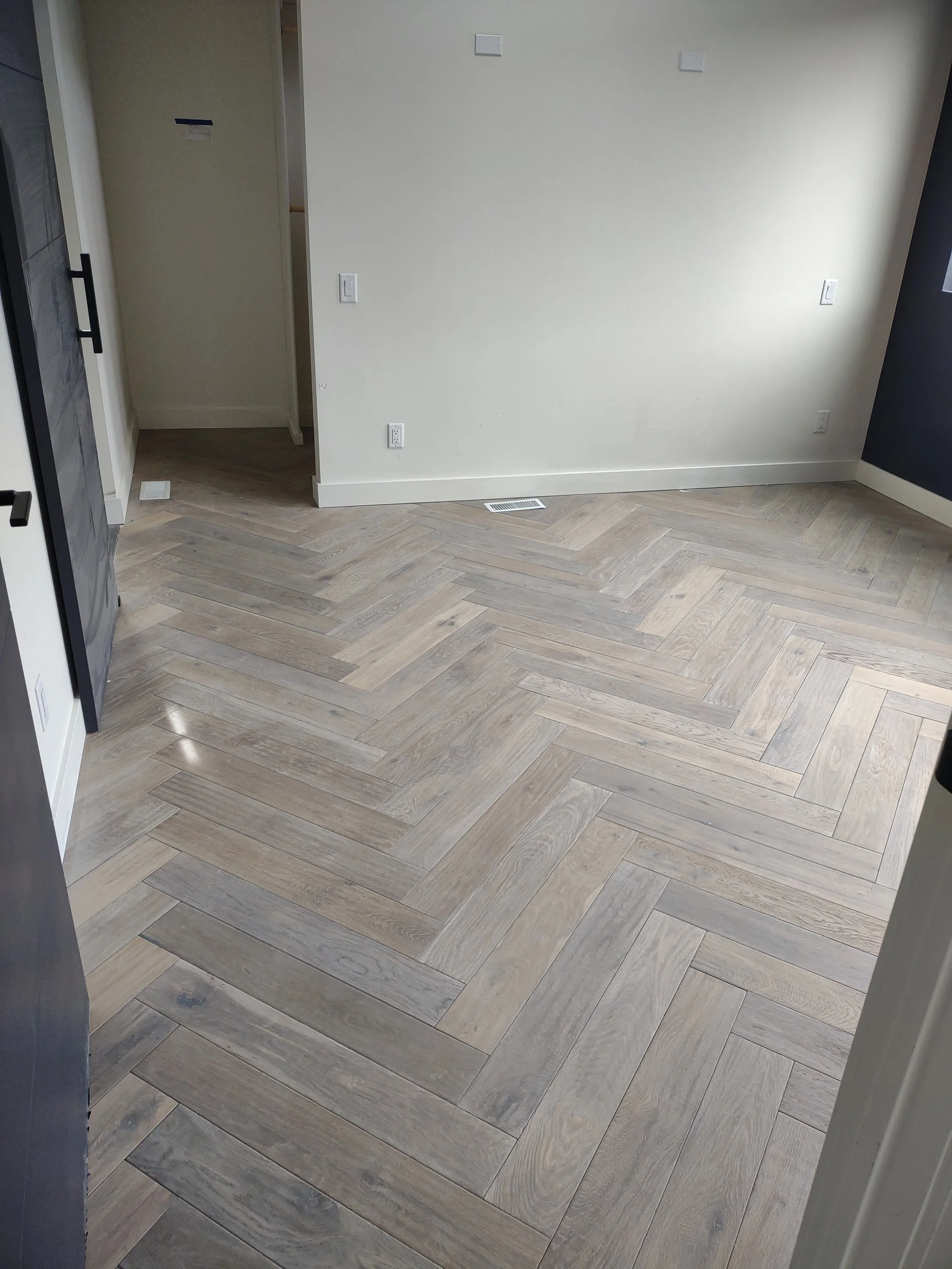Empty room with light-colored herringbone wood flooring, white and dark walls, and several electrical outlets.