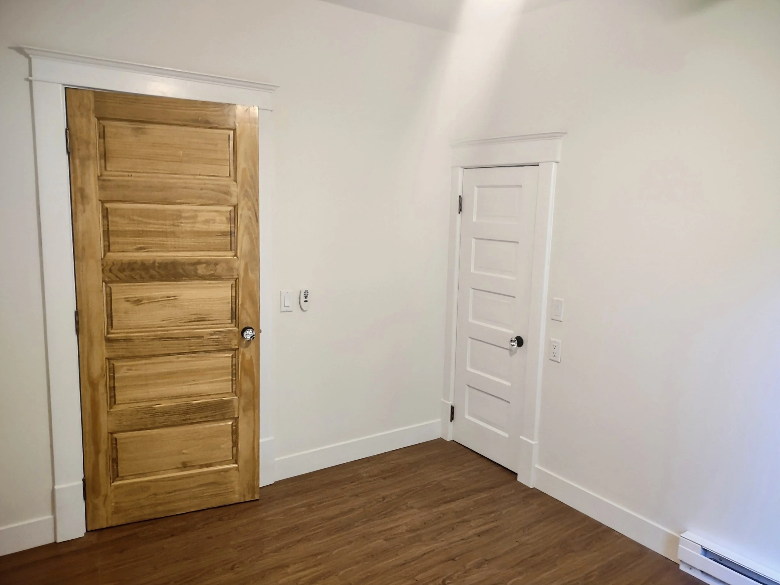 Empty room with two closed doors, one wooden and one white, with white trim and a small baseboard heater on the floor.