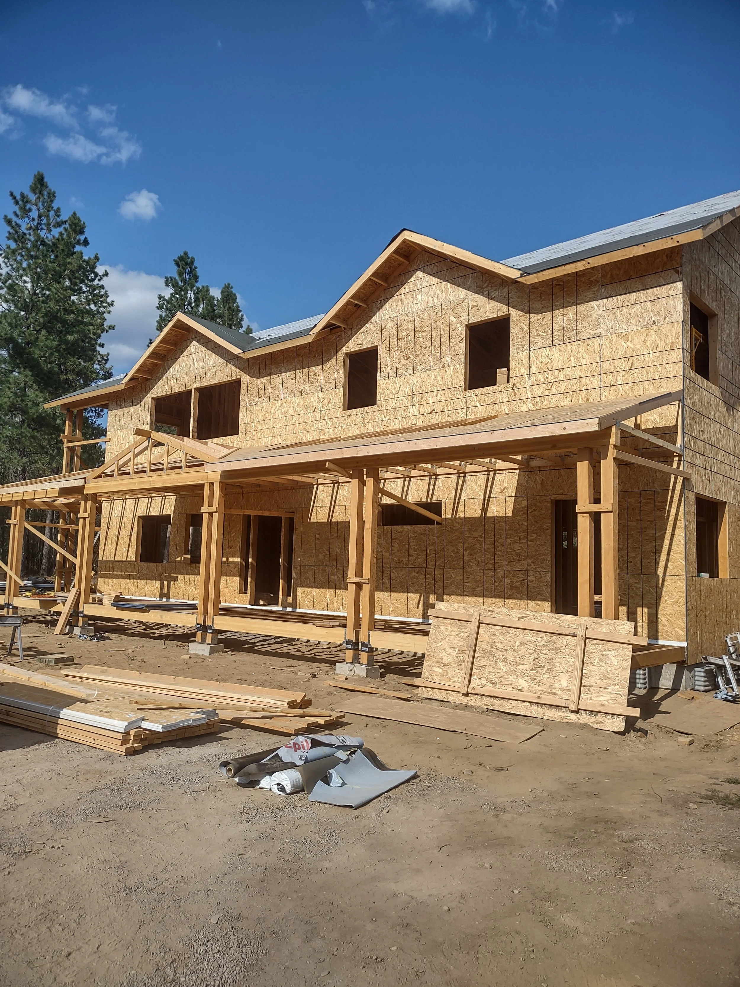 A house under construction with wooden framing and plywood sheathing, located in a wooded area on clear sunny day.