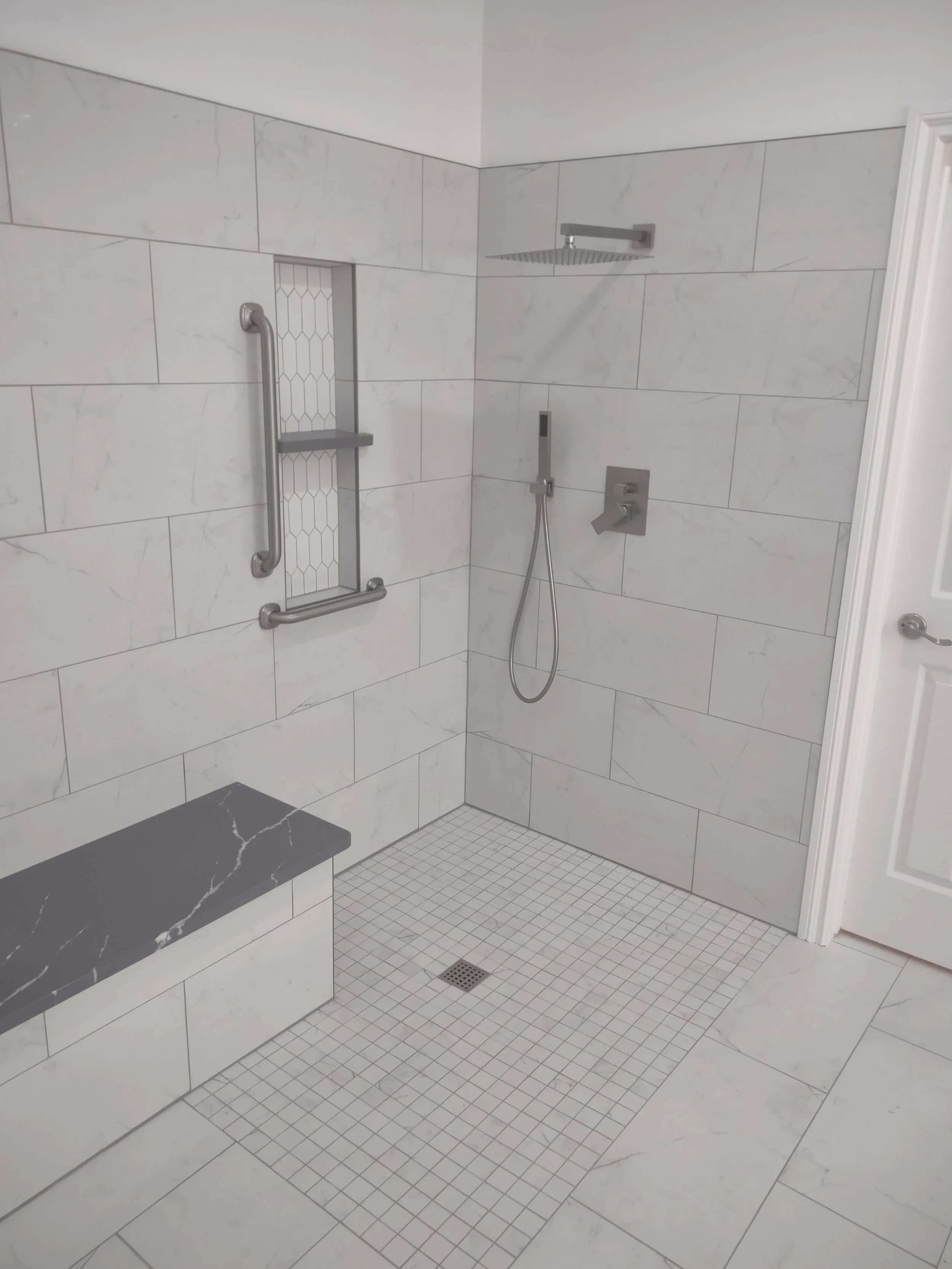 A modern, accessible walk-in shower with white marble tiles, a built-in bench with a black marble top, a handheld shower head, a rain shower head, and stainless steel grab bars