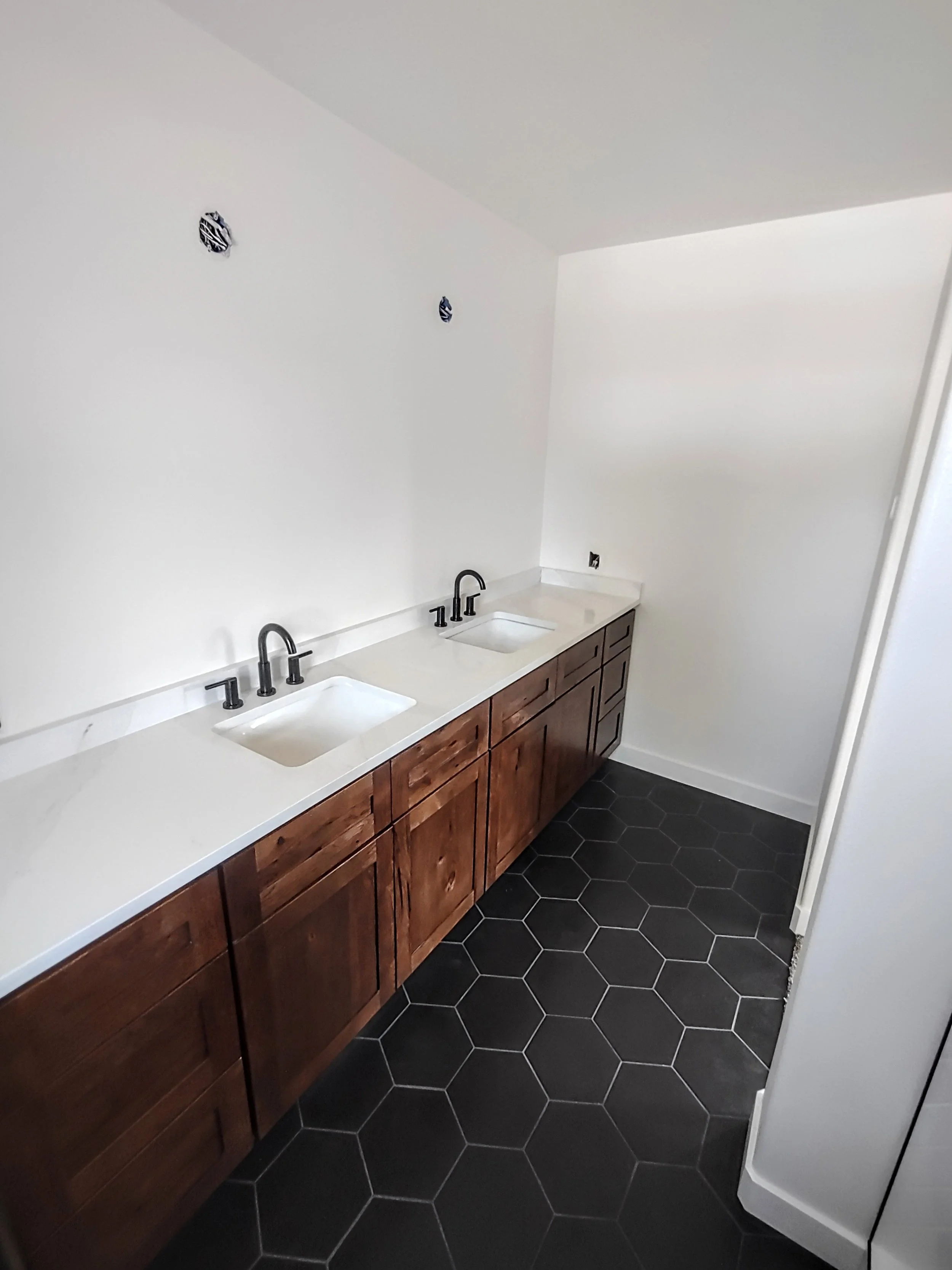 A bathroom vanity with two sinks, dark wood cabinets, black hexagonal floor tiles, and white walls.