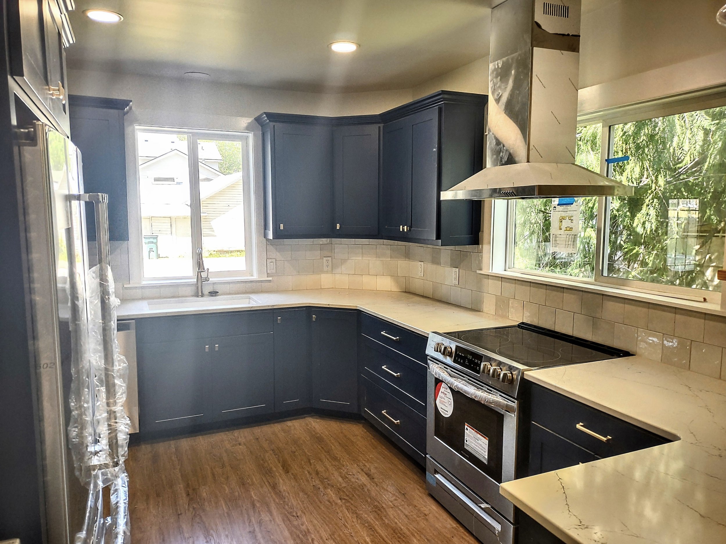 Modern kitchen with dark blue cabinets, beige countertops, a window over the sink, a stainless steel stove, a range hood, and hardwood flooring.