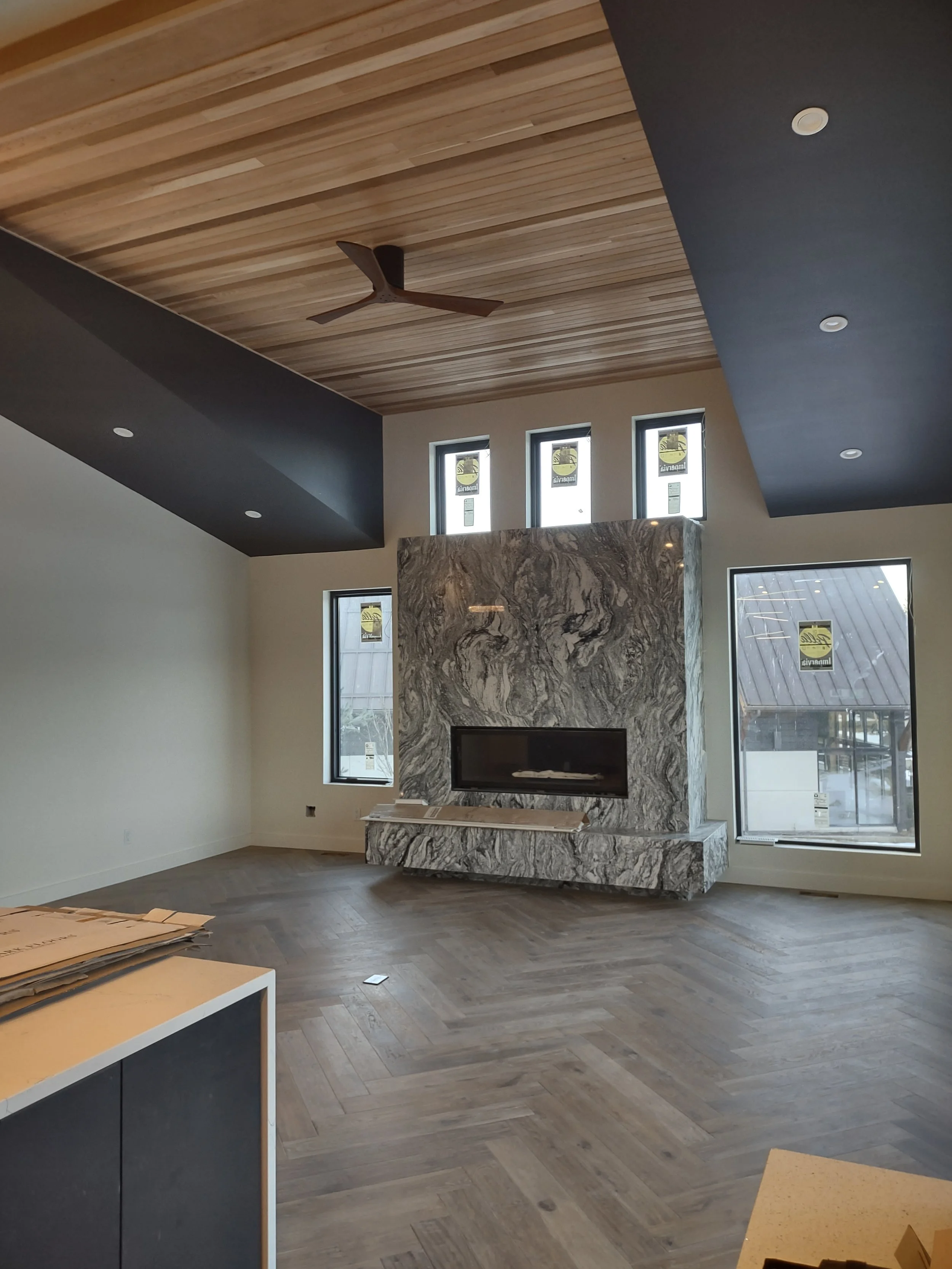 Modern living room with a wooden ceiling, black ceiling accents, a wood and gray marble fireplace, large windows, and light wood herringbone flooring.