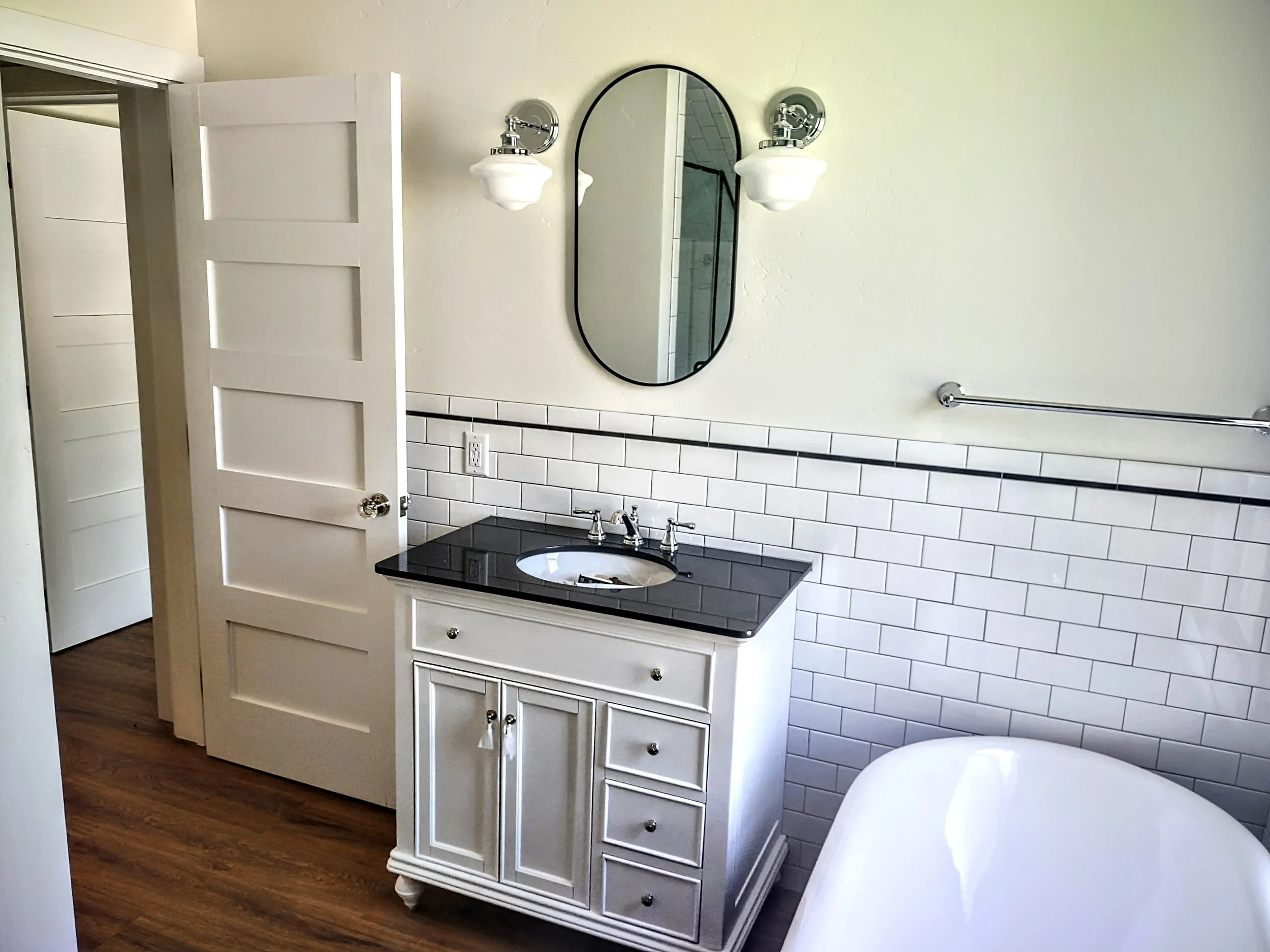 A bathroom with a white vanity with drawers and cabinets, a black countertop with a built-in sink, a mirror, two wall-mounted light fixtures, a towel rack, and a partial view of a bathtub. The wall has white subway tiles with a black border, and the 