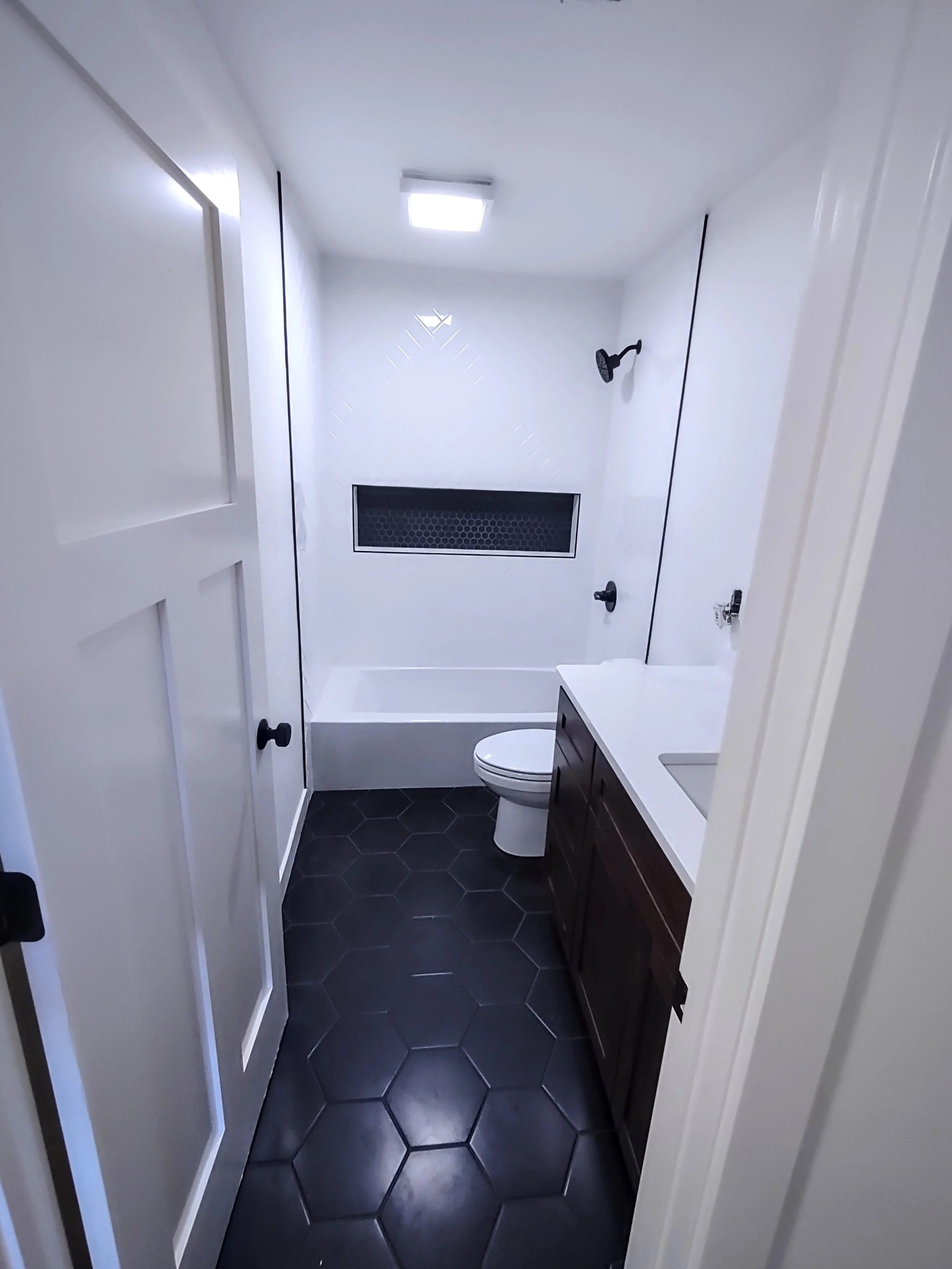 Small bathroom with a white shower area, black fixtures, dark hexagonal floor tiles, white walls, and a dark wooden vanity with a white countertop.