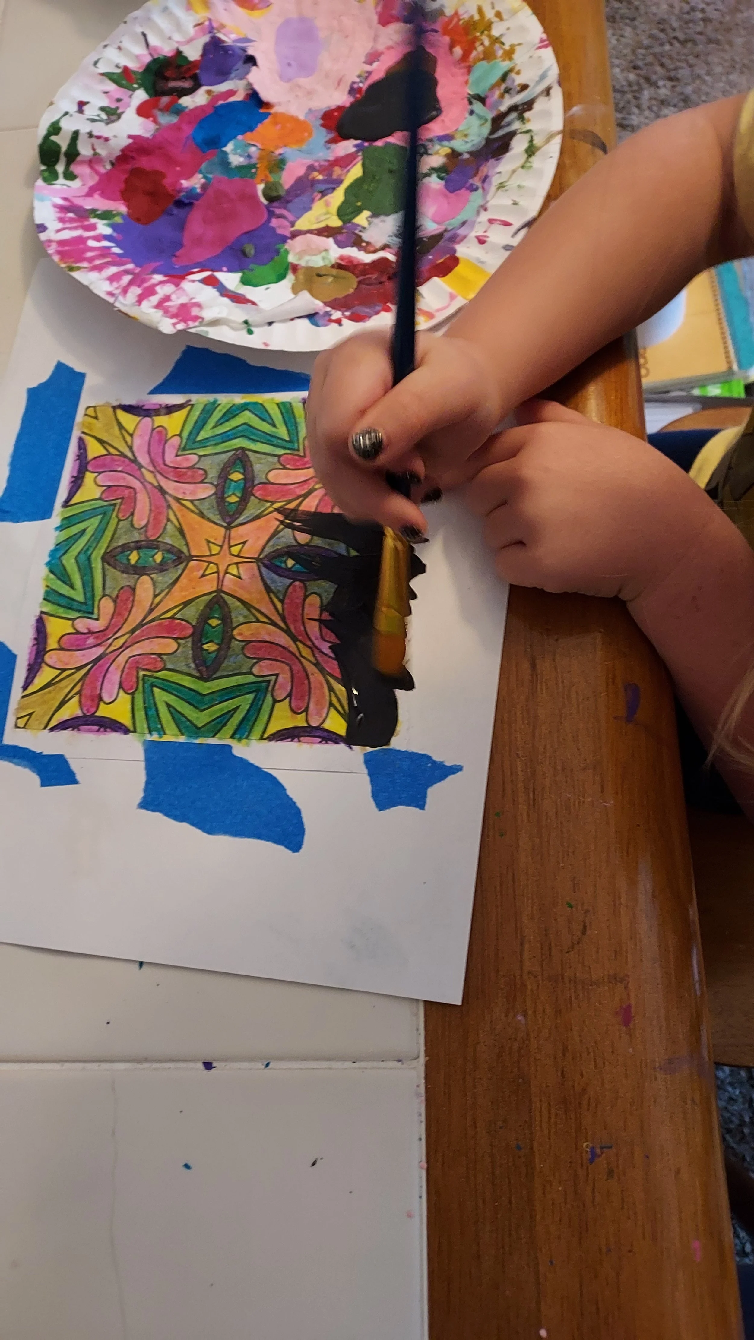 A child is coloring a detailed geometric and floral pattern on a piece of paper with crayons, sitting at a wooden table. There is a paint palette with mixed colors nearby.