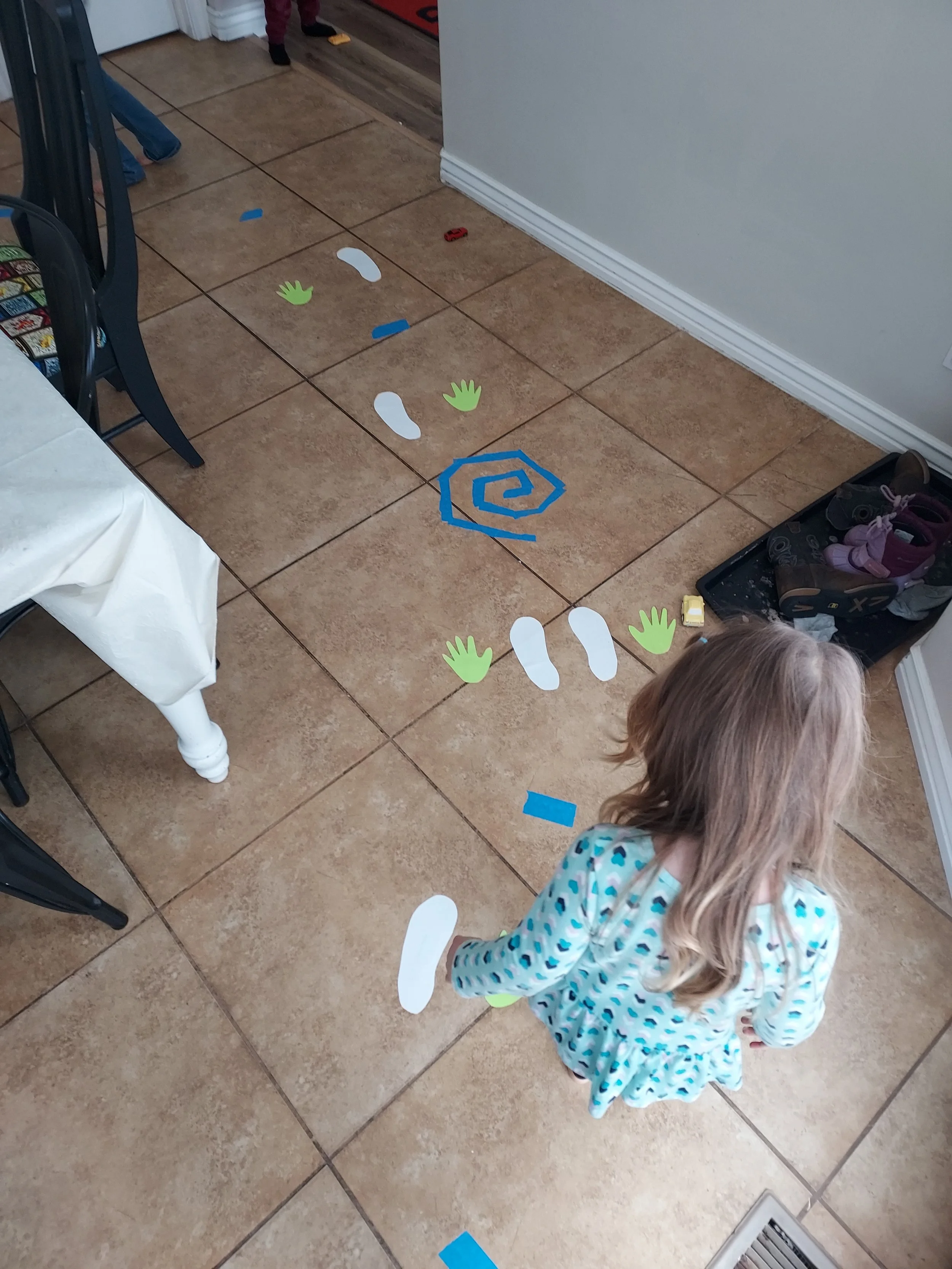 Child playing on tiled floor with paper cutout props including footprints, handprints, a spiral, and colored tape pieces.