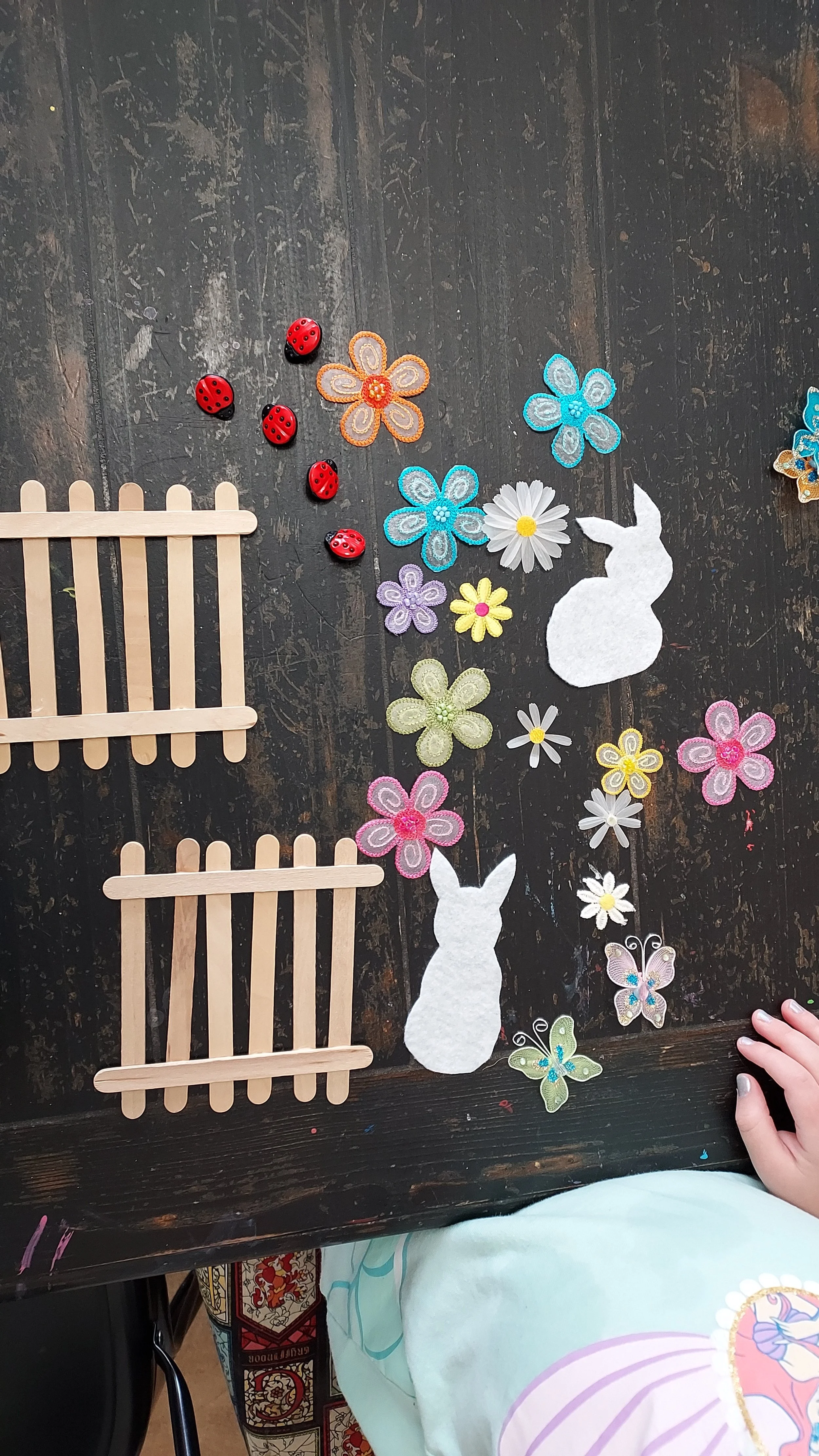 A flat lay of craft materials on a dark wooden surface, including felt bunnies, colorful felt flowers, small wooden fences, ladybug figurines, and a child's hand.
