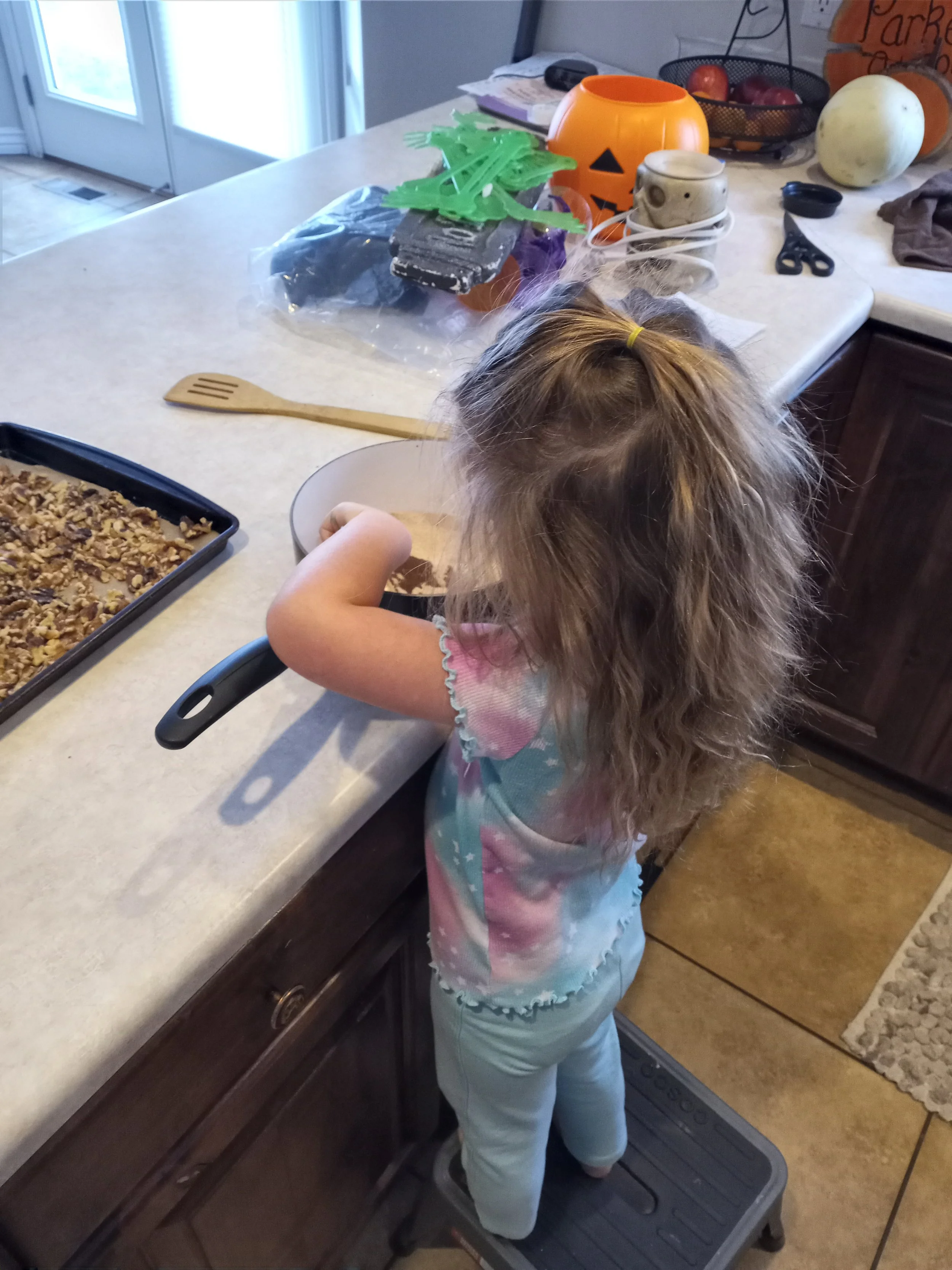 A young girl with long curly hair, wearing a colorful tie-dye shirt and white pants, is standing on a step stool while mixing ingredients in a large bowl in a kitchen. There is a baking tray with a dessert or casserole, various kitchen items, Hallowe