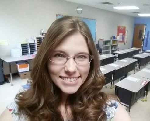 A smiling teacher with glasses taking a selfie in a classroom with desks and shelves in the background.