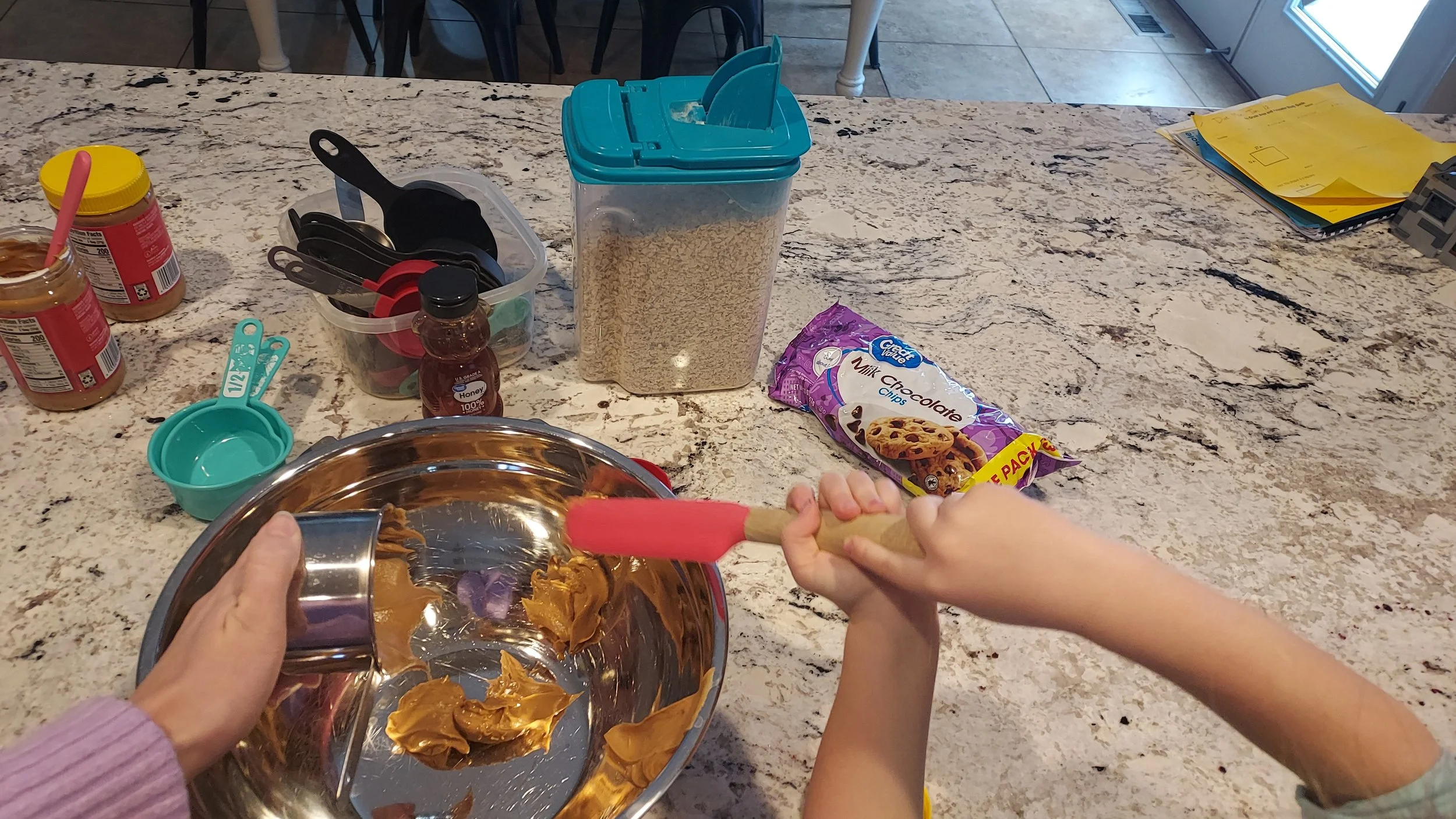 Child spreading peanut butter on a knife over a stainless steel mixing bowl, with peanut butter inside. Various baking ingredients and utensils are scattered on a granite kitchen countertop.