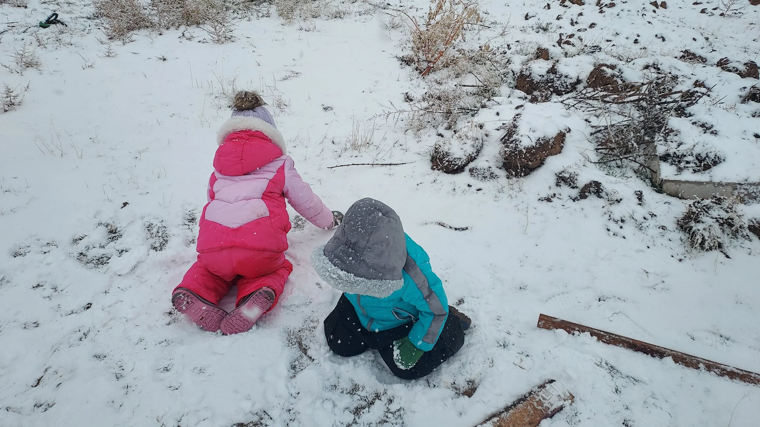 Two children wearing winter coats, hats, and gloves playing in the snow outdoors, kneeling near a small snow pile.