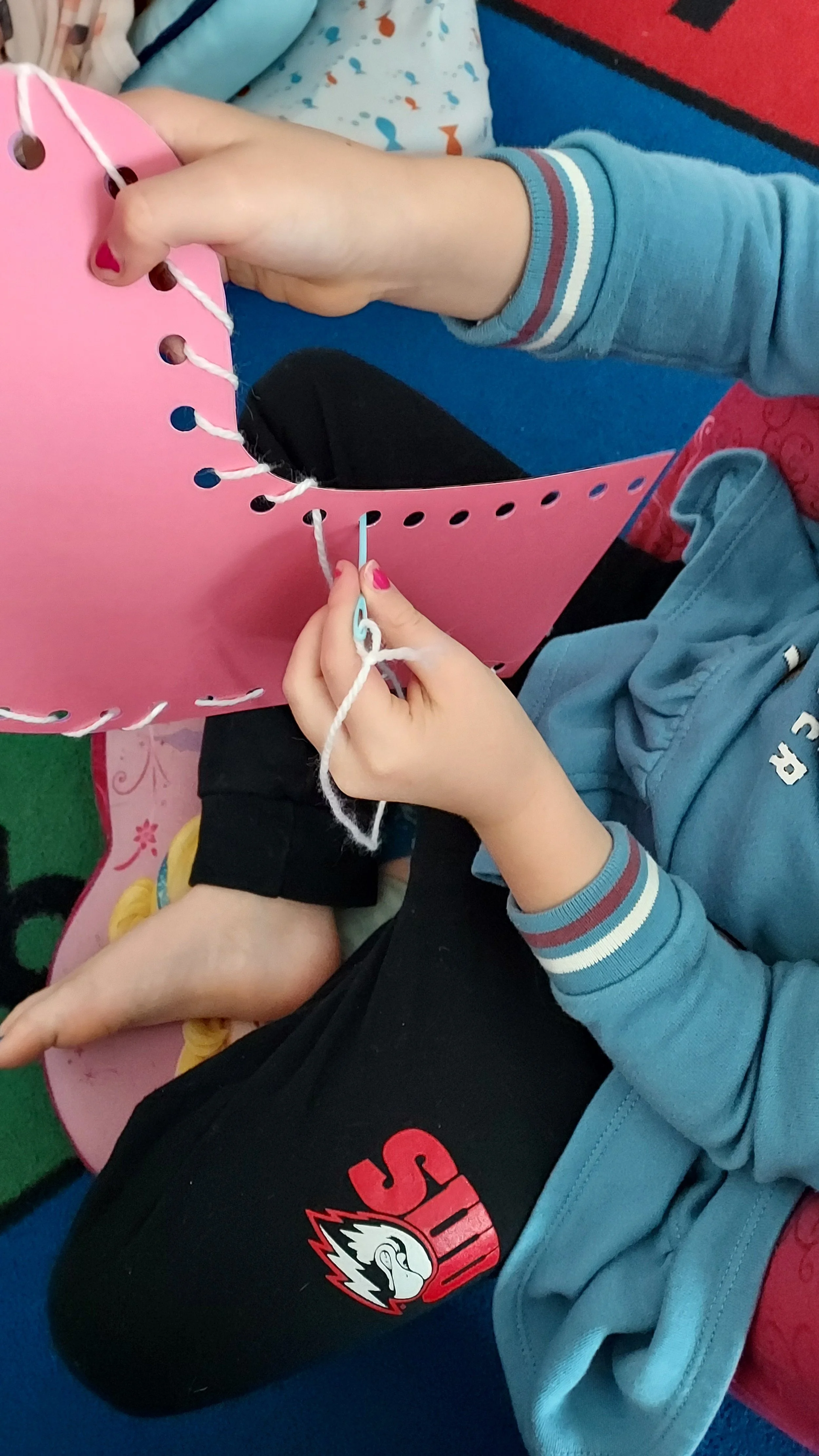 A person is sewing or lacing a pink cardboard or paper craft, using a white string and a needle. The person is wearing a blue sweatshirt with striped cuffs. There are children with their legs crossed sitting nearby on a blue and red carpet.