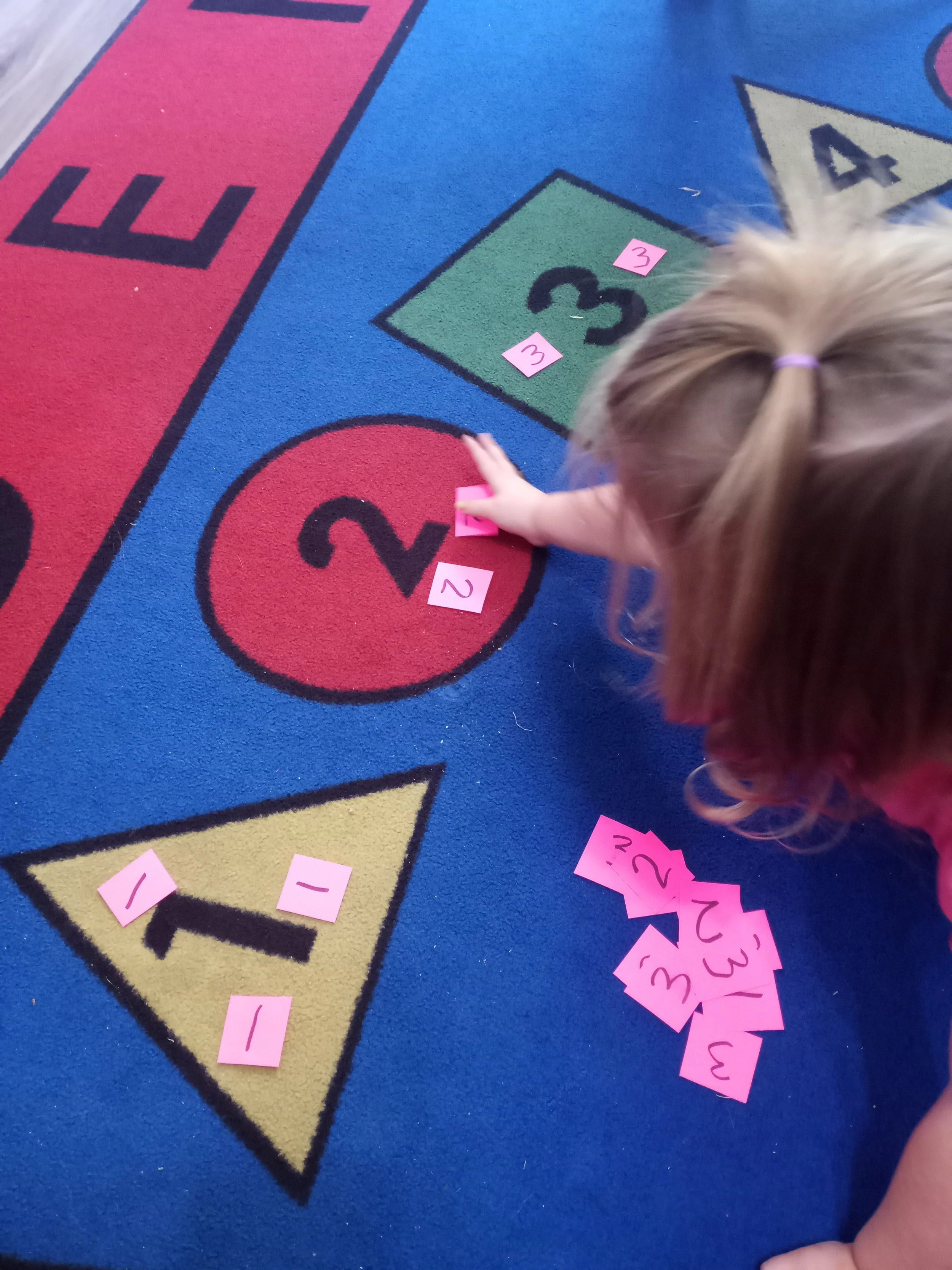 A child is placing pink number cards on a colorful educational game rug that features numbers and shapes, with some cards on the floor.