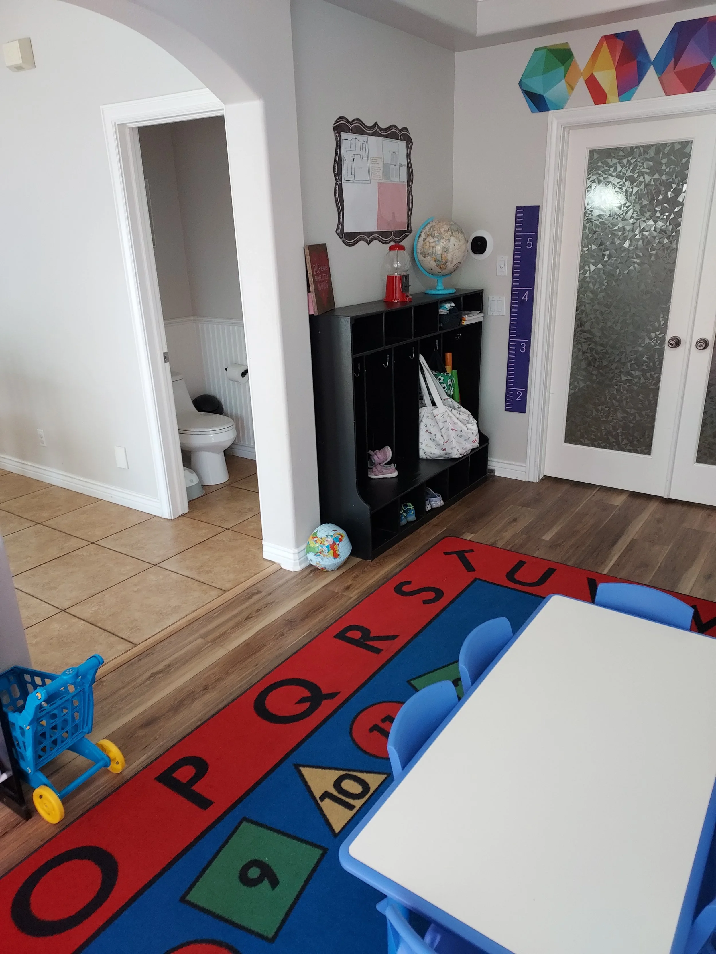 Colorful classroom corner with a black cubby shelf holding books and a globe, a purple growth chart on the wall, a white table with blue chairs, a red educational rug with letters and numbers, and a small grocery cart toy on the floor.
