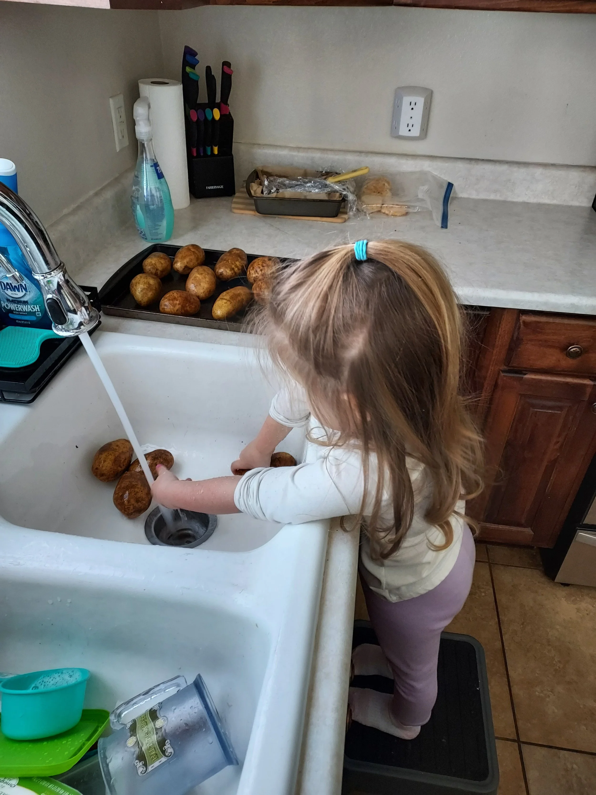 A young girl with long hair is washing potatoes in a kitchen sink.